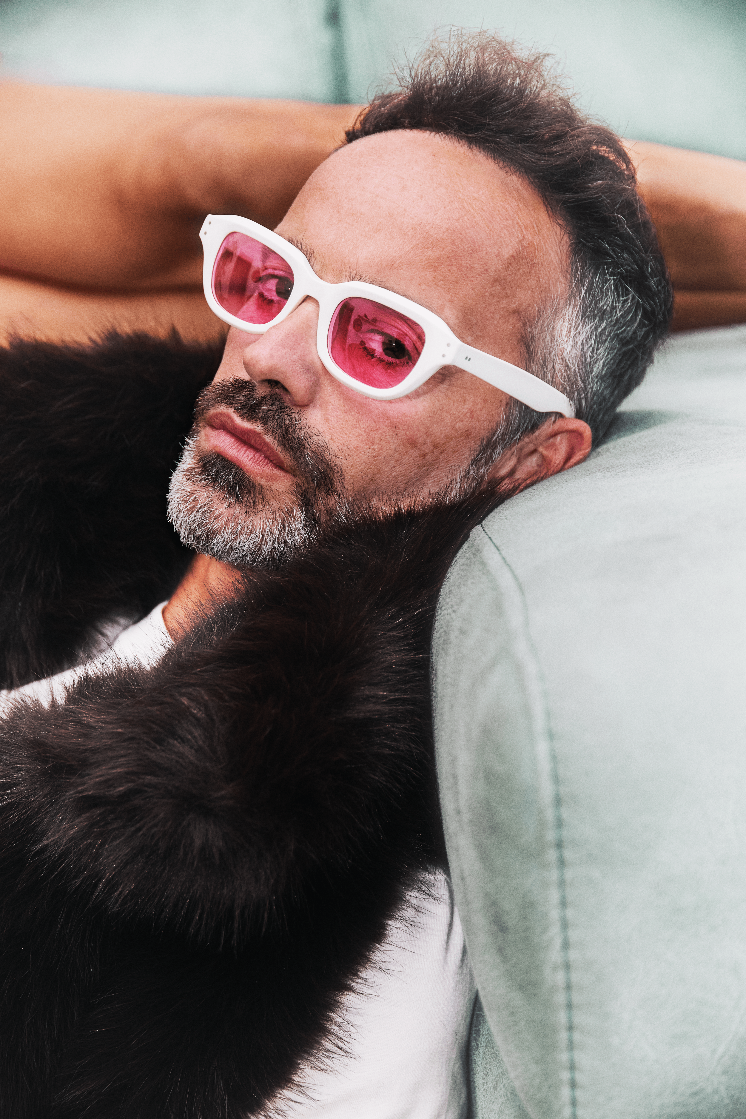 A man with gray facial hair and black hair reclining on a sofa. He is wearing white sunglasses with pink lenses and a black furry coat.
