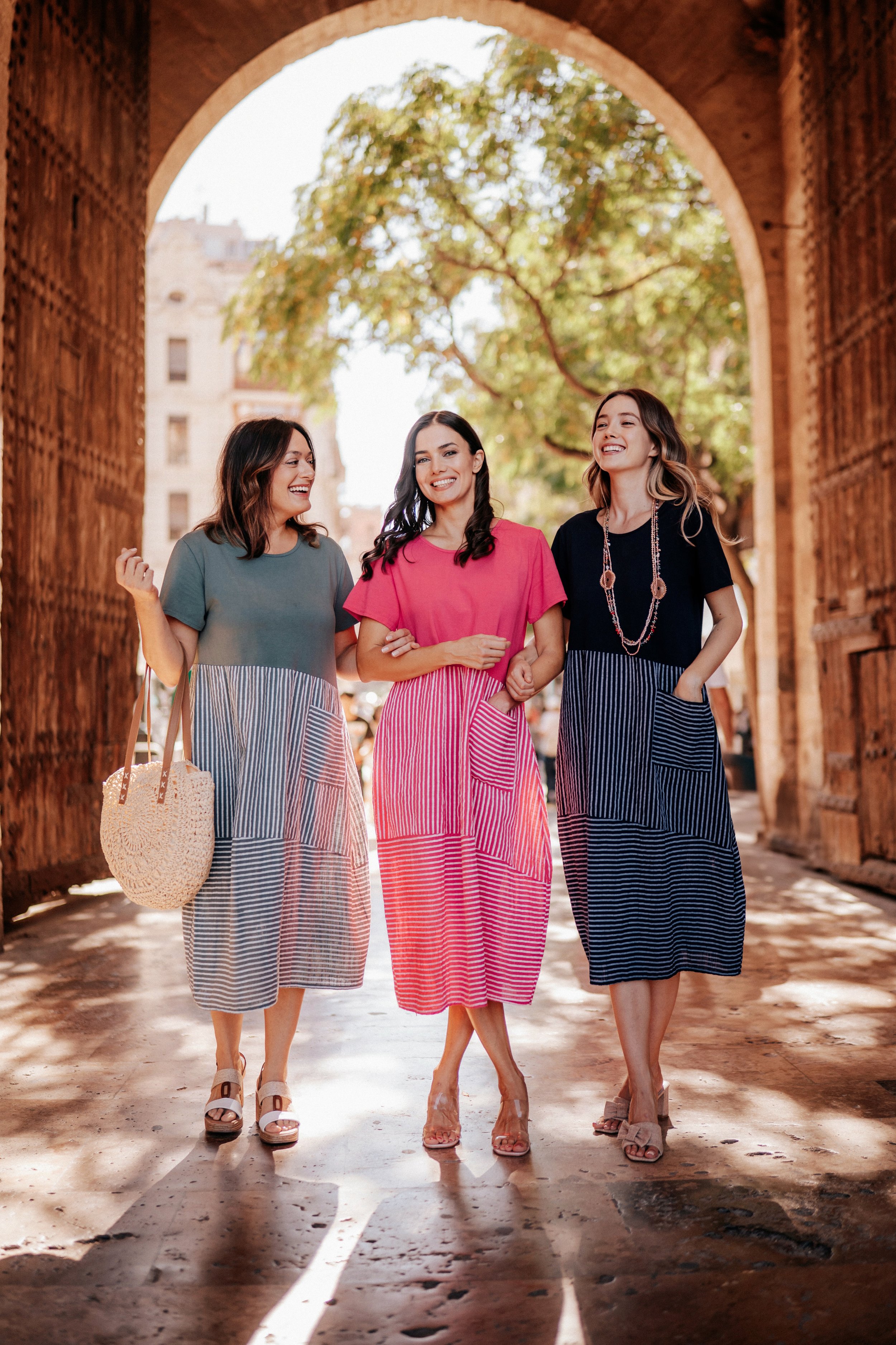 Three women walking together under an archway, smiling and talking, in an urban setting with trees and buildings in the background.