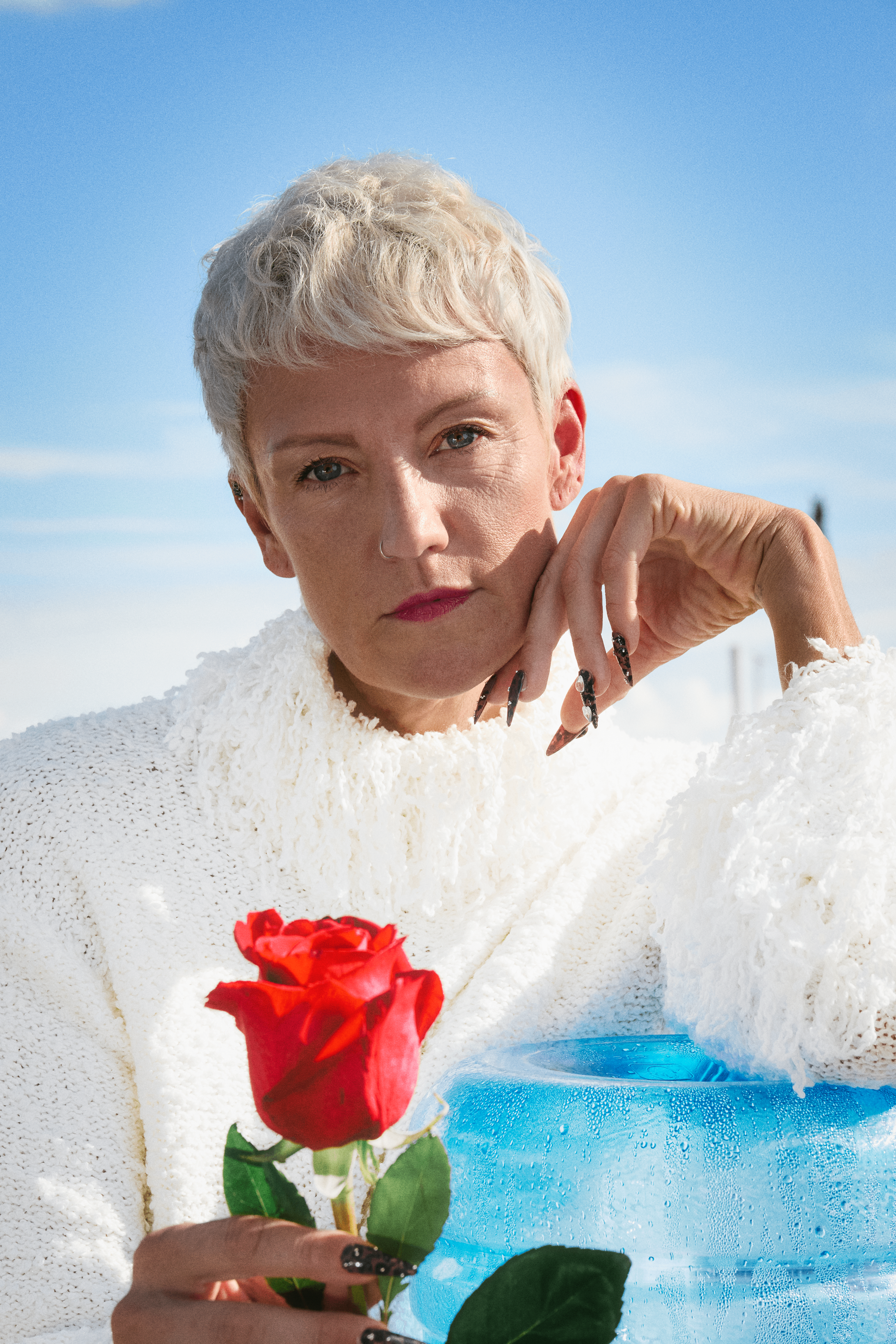 A woman with short platinum blonde hair, wearing a white sweater, holding a red rose, and resting her face on her hand against a blue sky background.