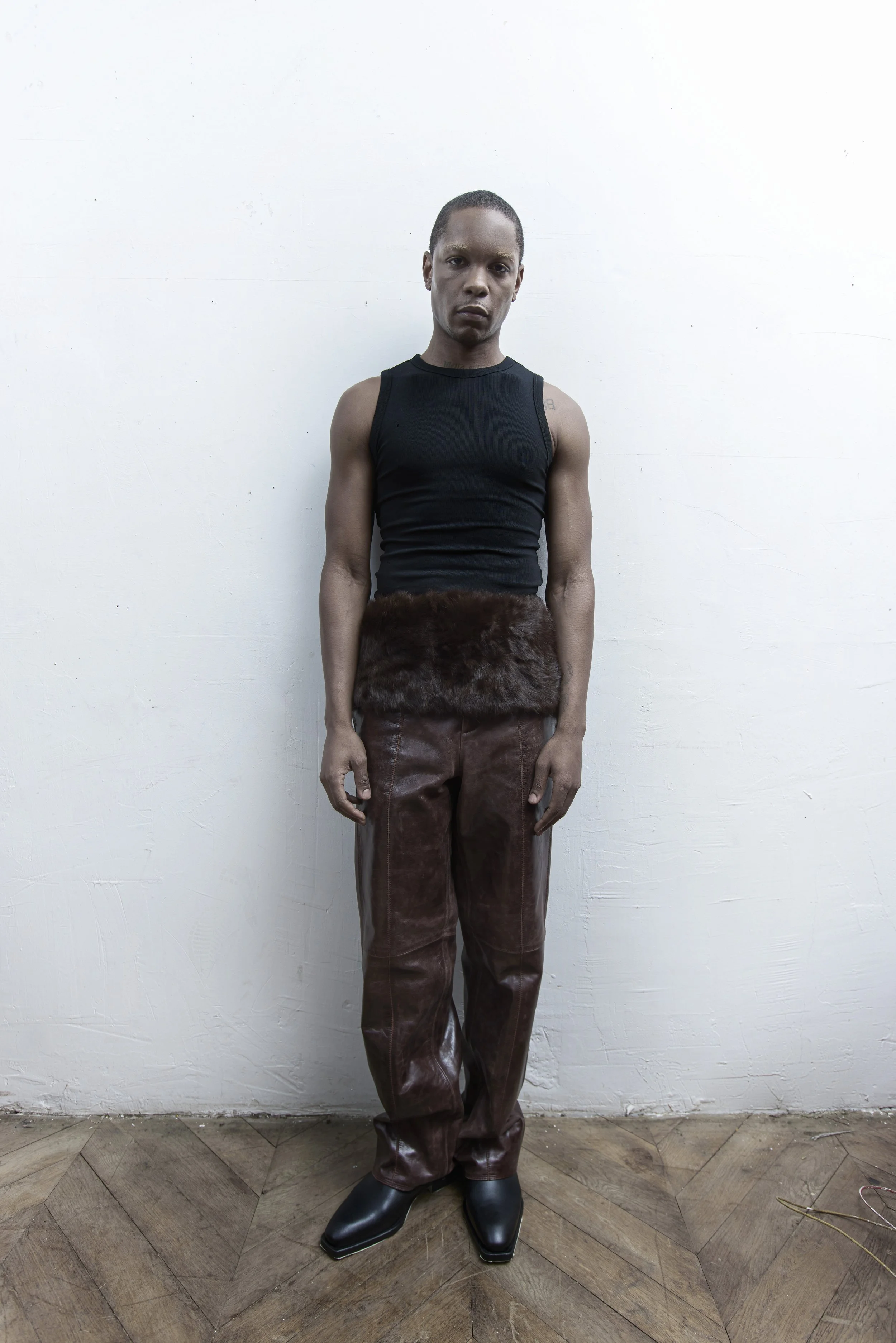 Man standing against a white wall, wearing a black sleeveless top, brown leather pants with fur waist detail, and black boots.