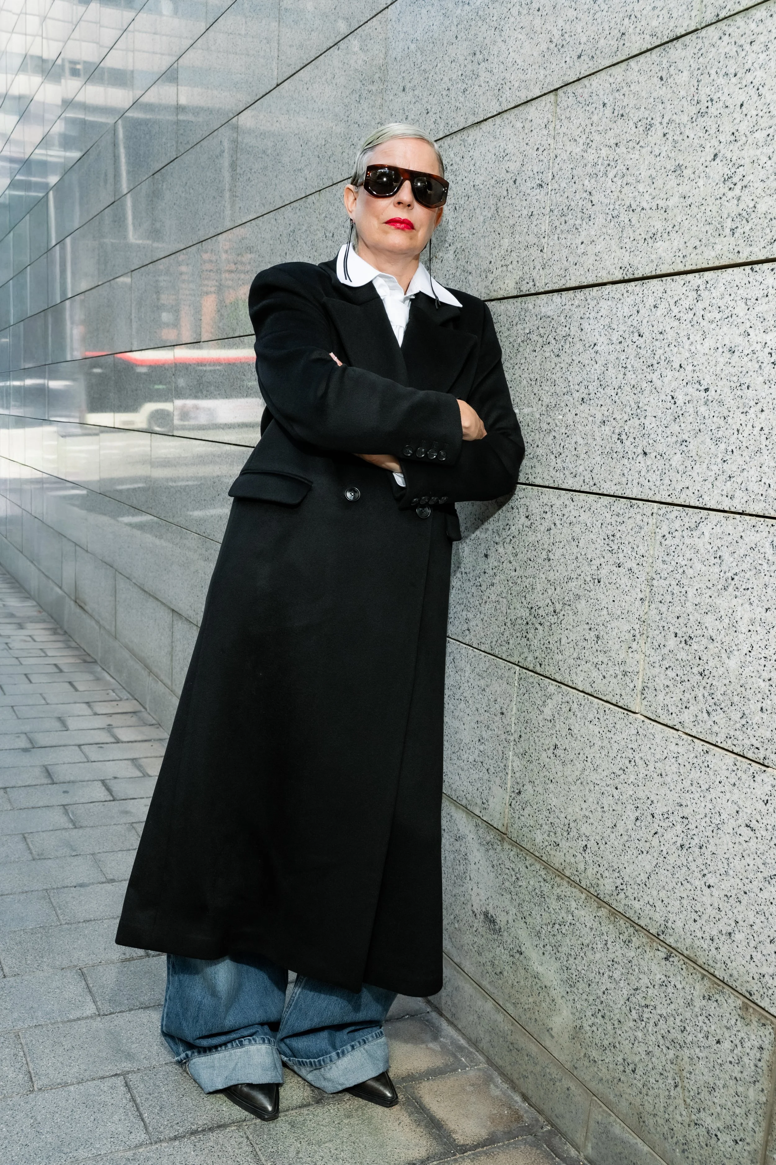 A woman with gray hair wearing large sunglasses, a white collared shirt, a black blazer, a long black skirt, and baggy blue jeans, standing with her arms crossed and leaning against a speckled gray stone wall on a city sidewalk.