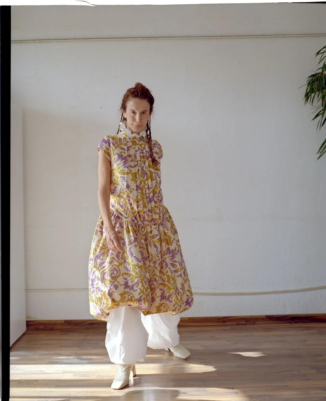 A woman in a colorful, patterned dress with yellow and purple accents, white puffy pants, and white boots standing on a wooden floor in front of a plain white wall.