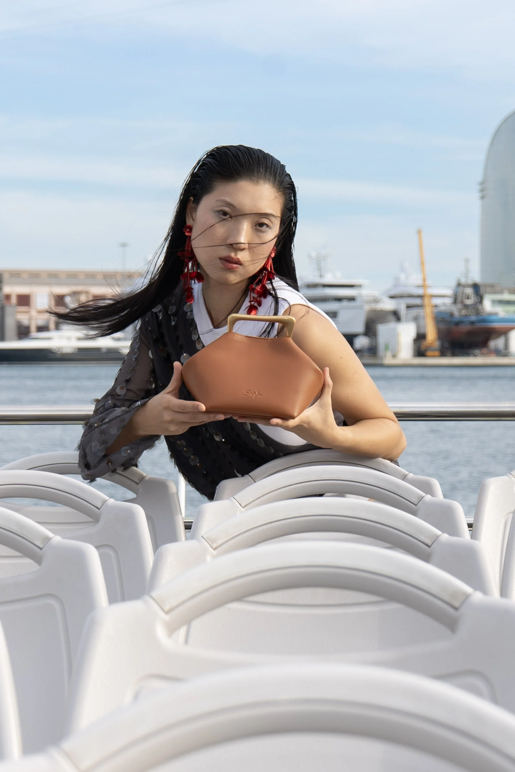 A woman with long black hair, wearing red earrings and a black and white outfit, is sitting outdoors near water, holding a brown handbag, with boats and buildings in the background.