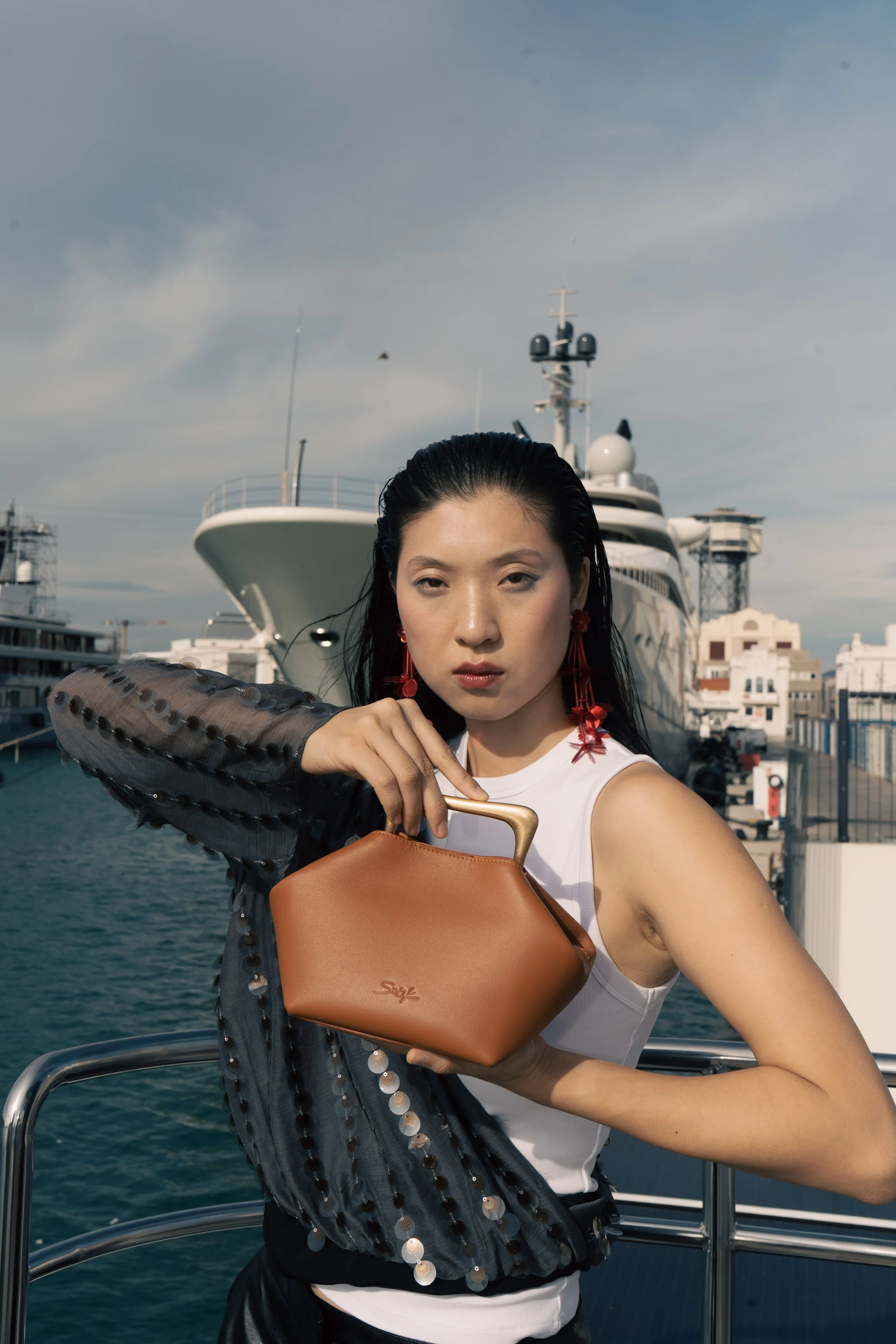 A woman with wet black hair is holding a tan handbag and posing on a yacht dock with a large yacht in the background under a partly cloudy sky.
