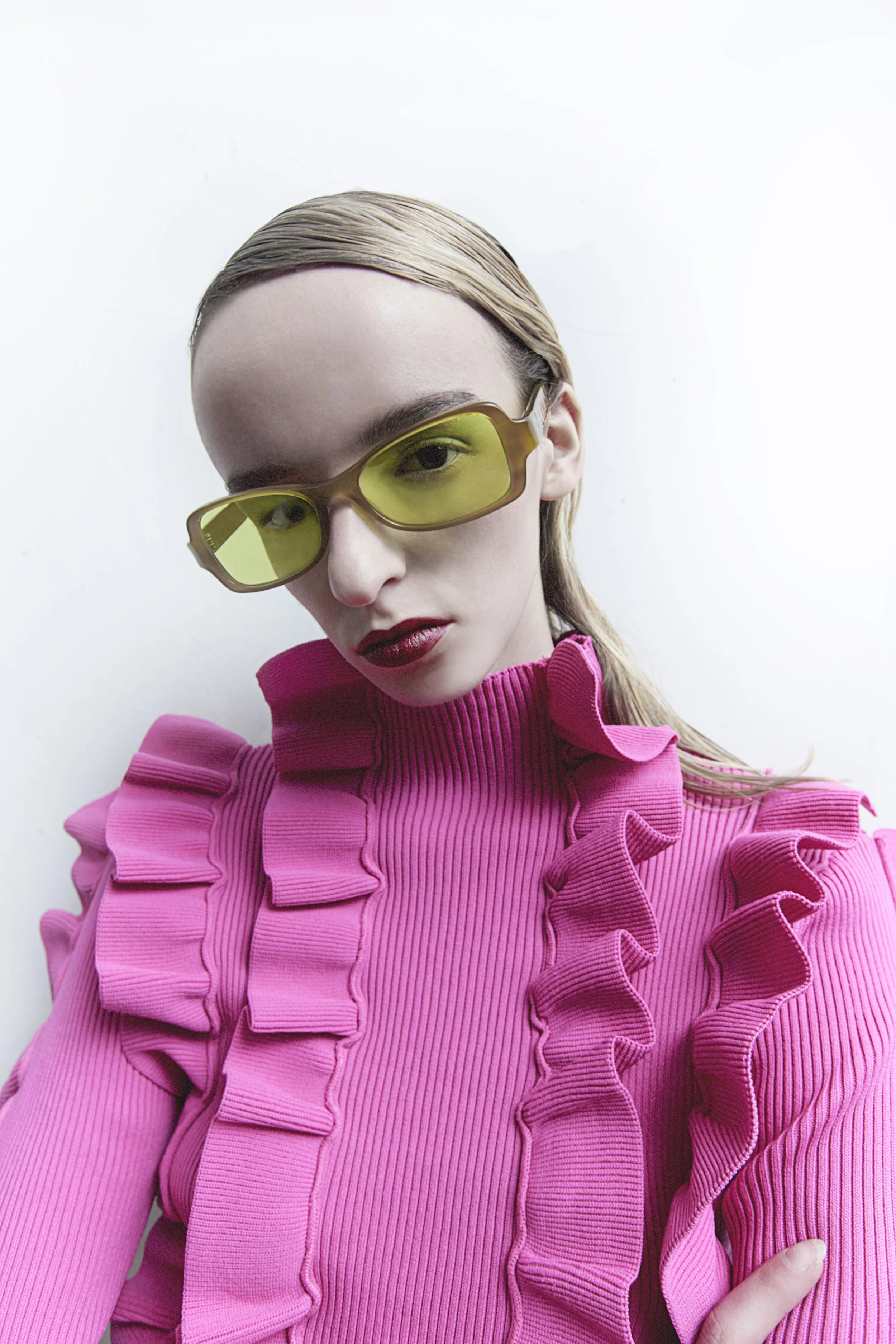 A woman with blonde hair wearing yellow-tinted sunglasses and a bright pink ruffled top, posing against a plain white background.