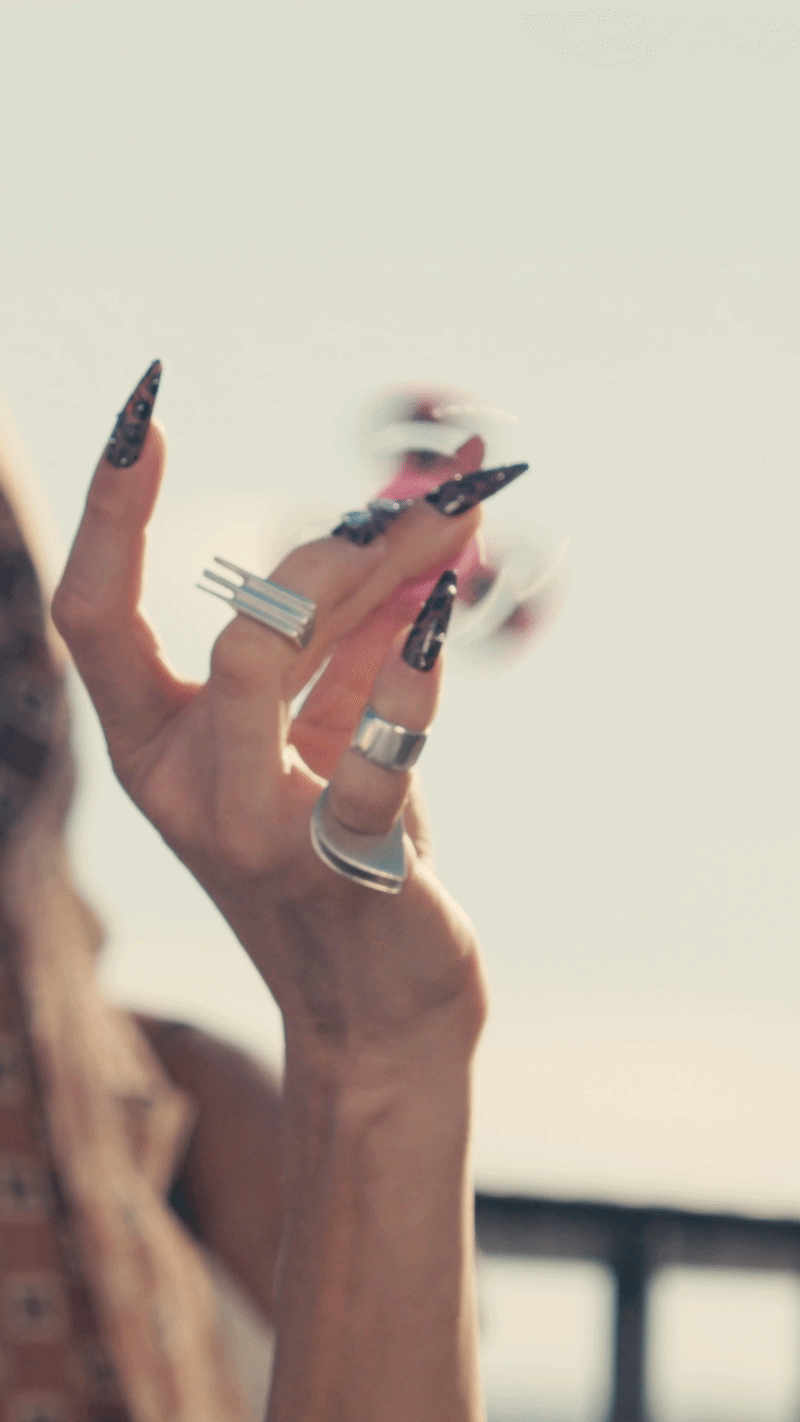 Close-up of person's hand with long black nails and silver rings, holding and blowing smoke from a cigarette.