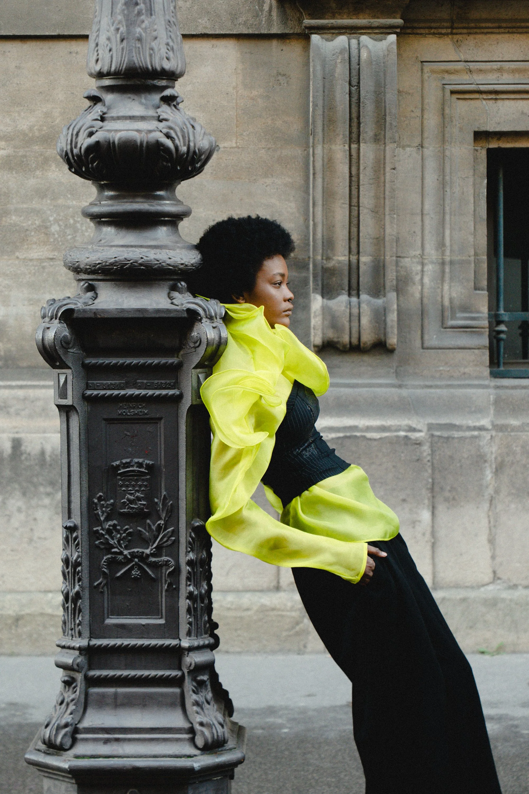 A woman with natural hair wearing a yellow blouse and black dress, leaning against a decorative streetlamp in front of a stone building.