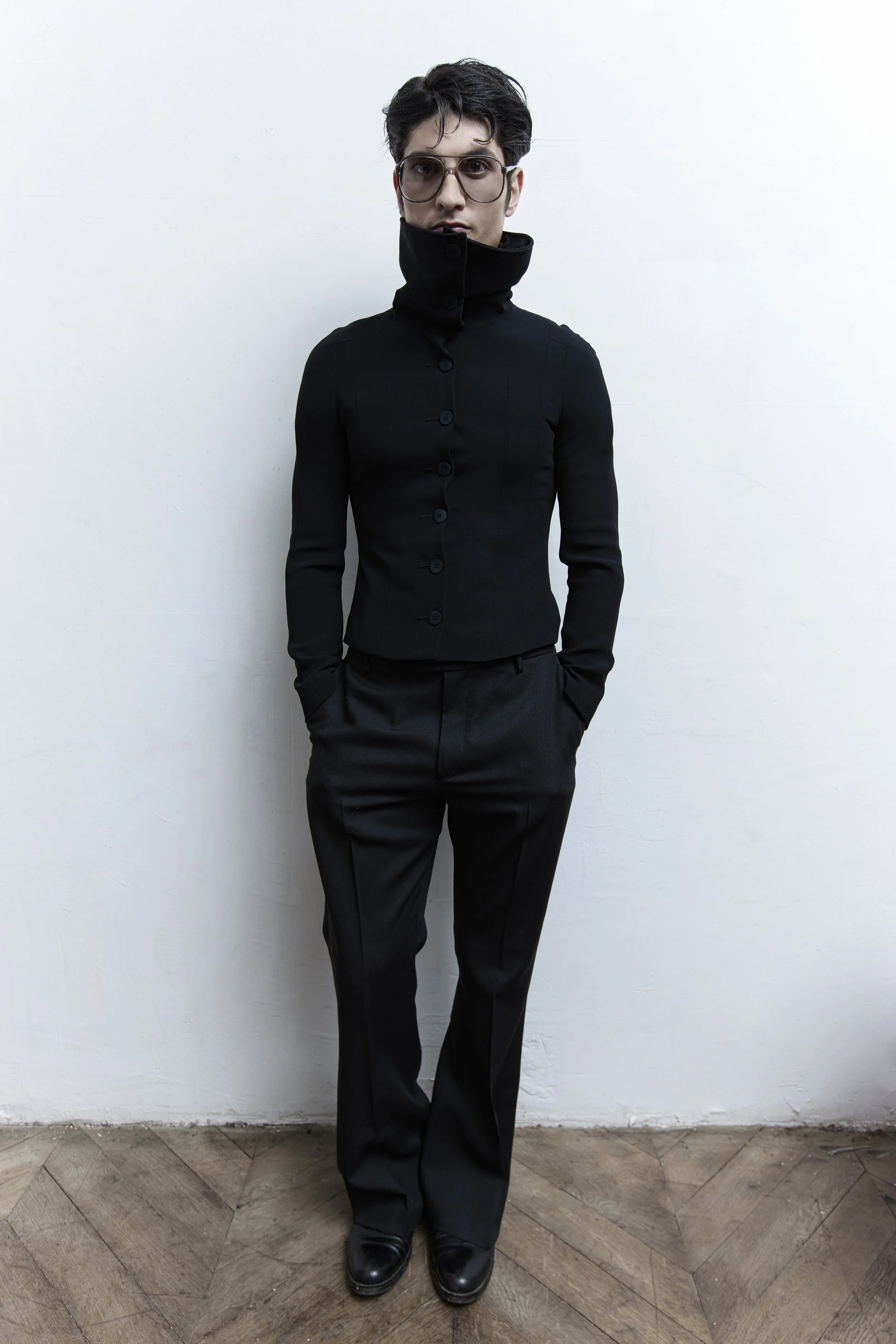 A person with dark, curly hair wearing sunglasses, a black high-collared jacket, black pants, and black shoes, standing against a plain white wall.