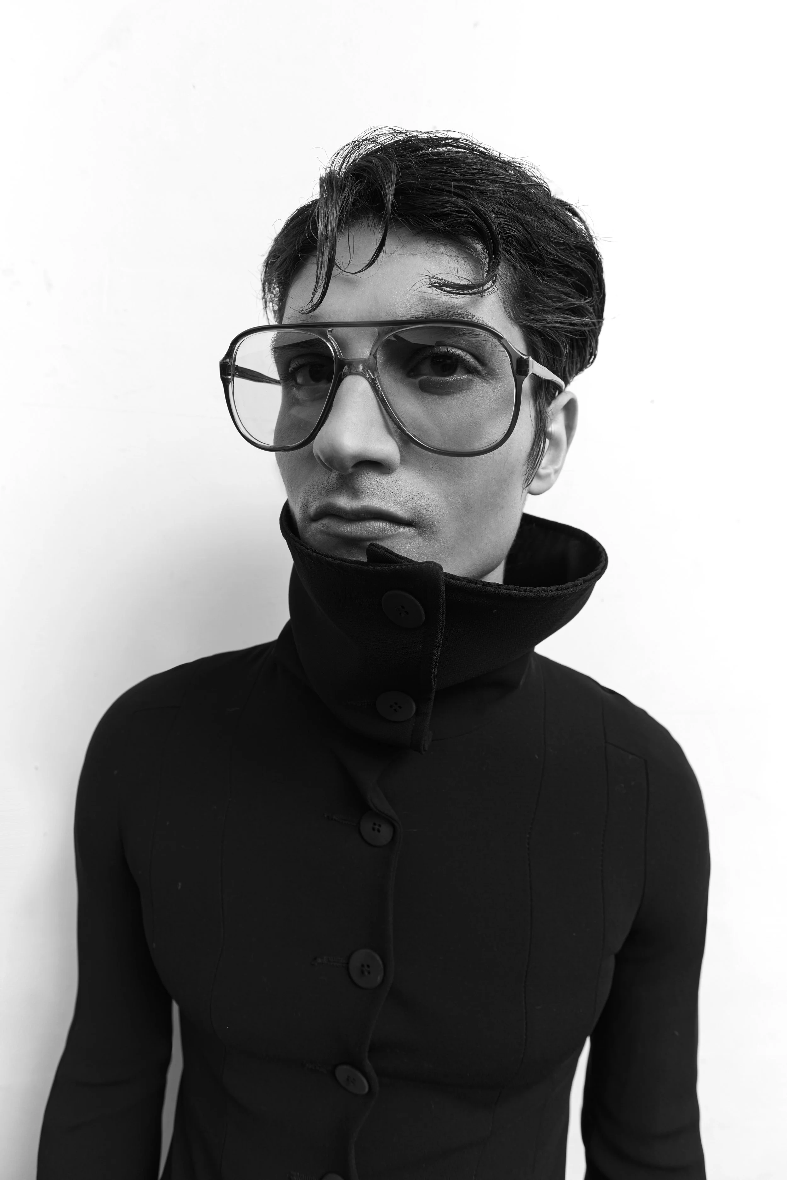 A black and white photograph of a person with short hair, wearing large glasses and a high-collared black coat, standing against a light background.
