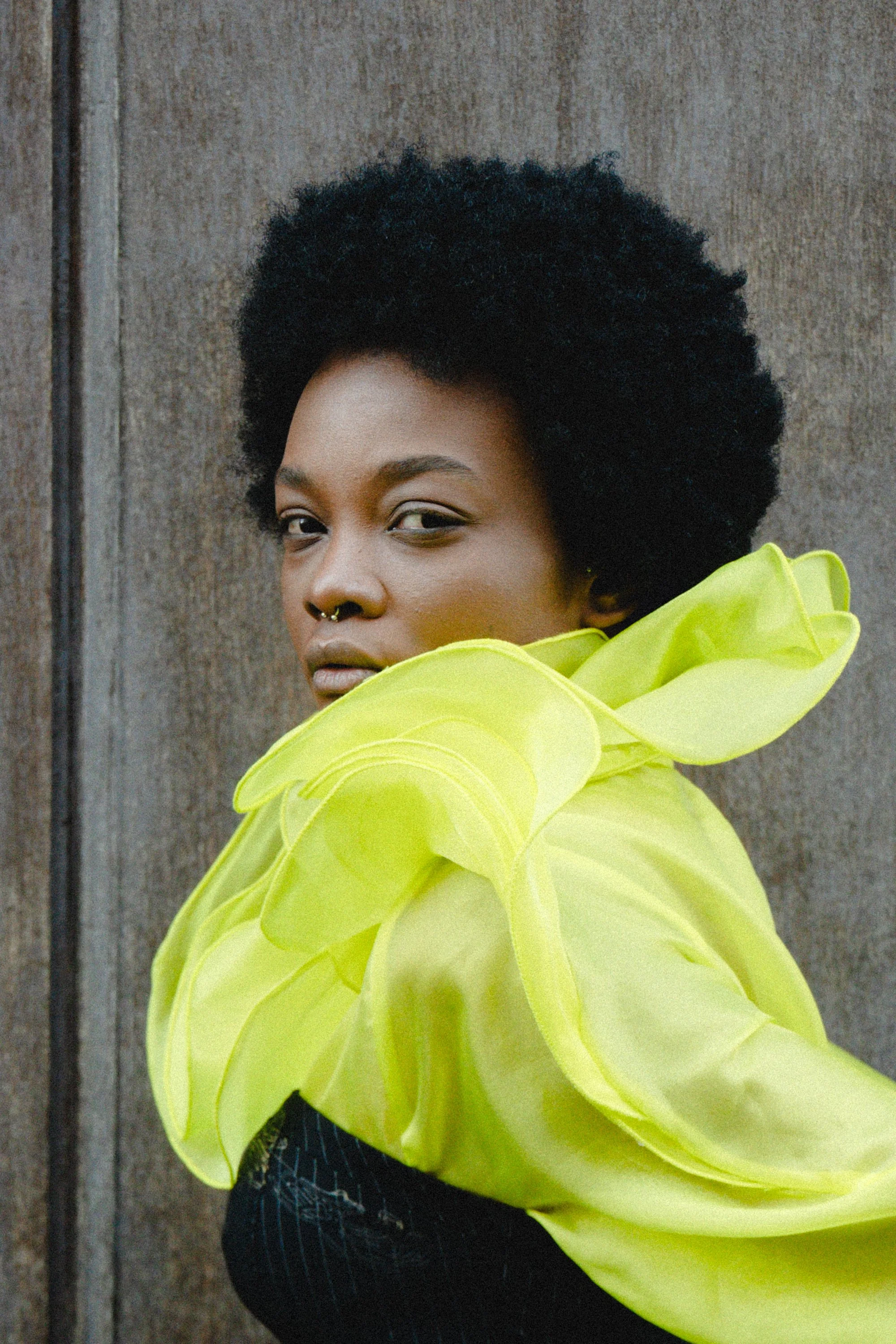 A woman with dark skin, short natural hair, and a septum piercing looking at the camera, wearing a bright yellow, sheer, ruffled garment over a black top, standing in front of a wooden background.