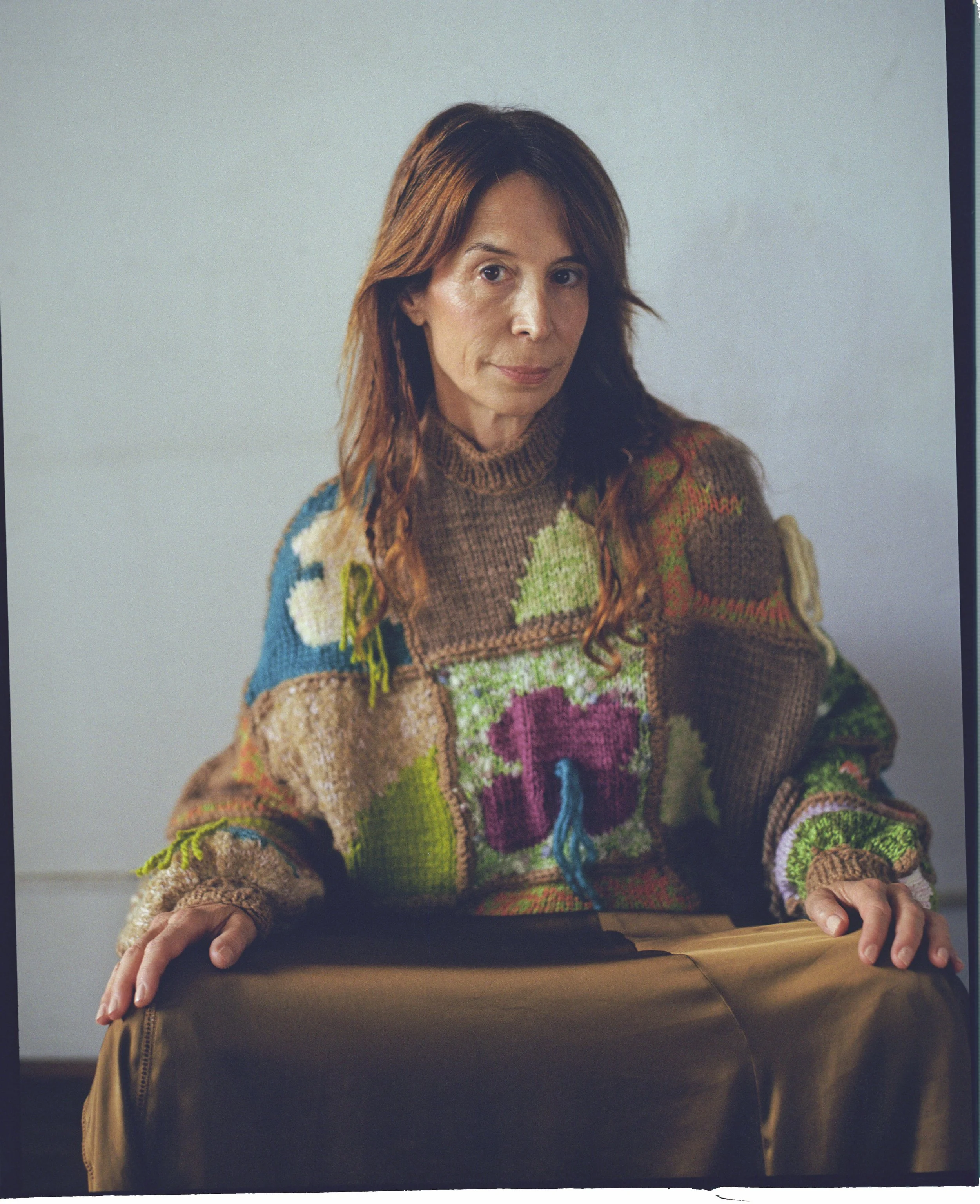 A woman with long reddish-brown hair wearing a colorful patchwork sweater sitting at a table.