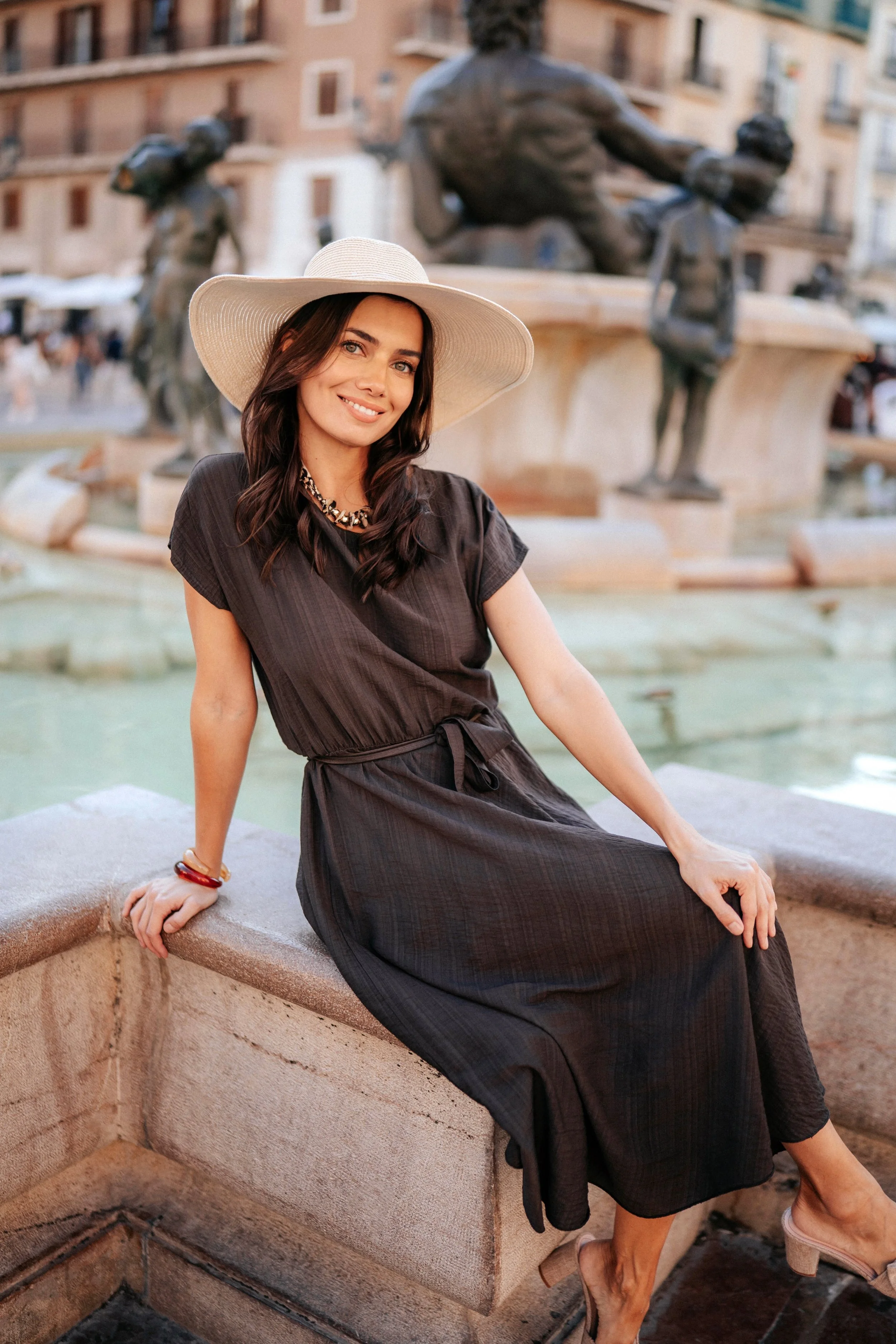 A woman sitting on the edge of a fountain in a city square, wearing a black dress and a wide-brimmed hat, with a sculpture and buildings in the background.