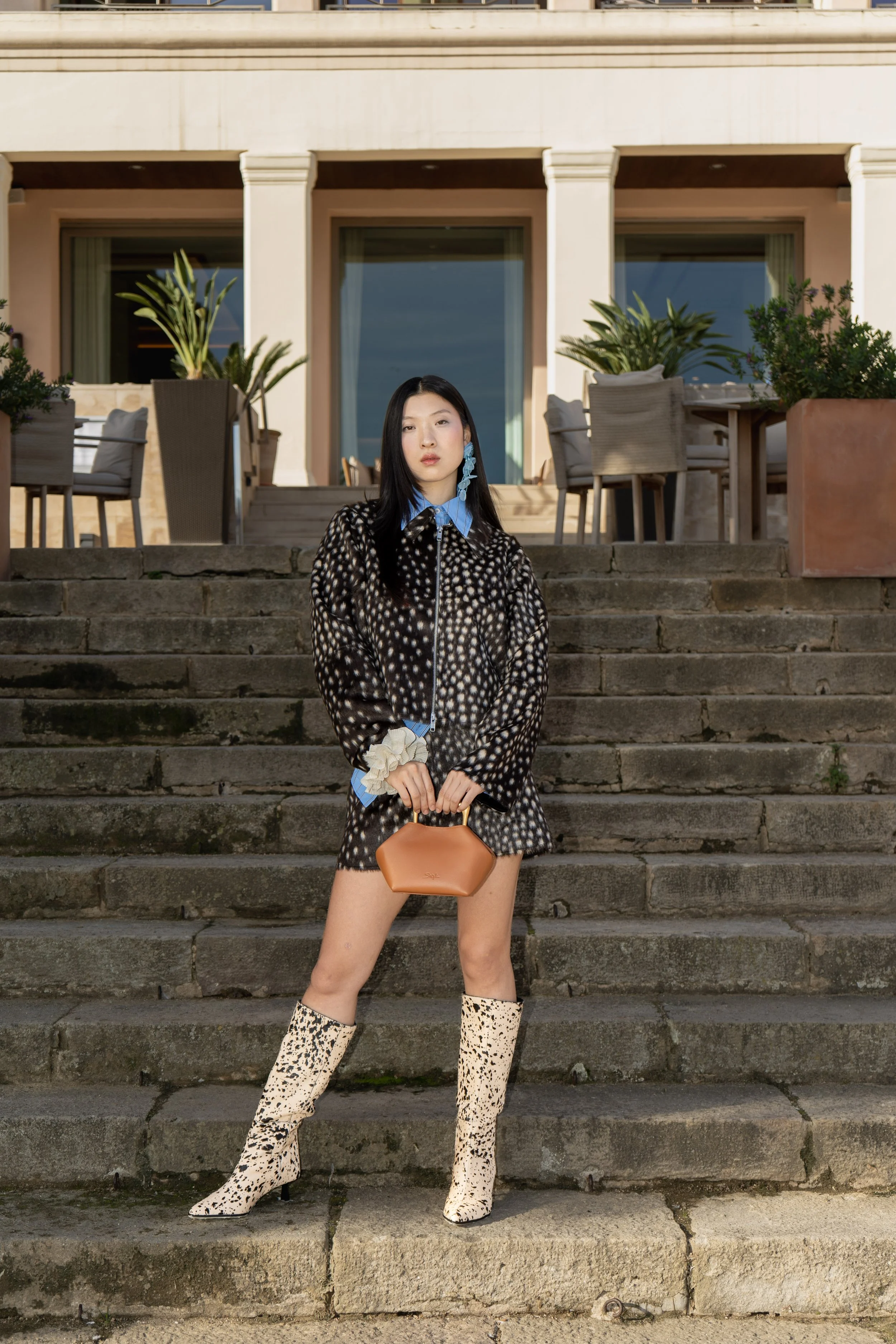 A woman in a patterned jacket and knee-high leopard print boots standing on stone steps outside a building with patio furniture and potted plants.