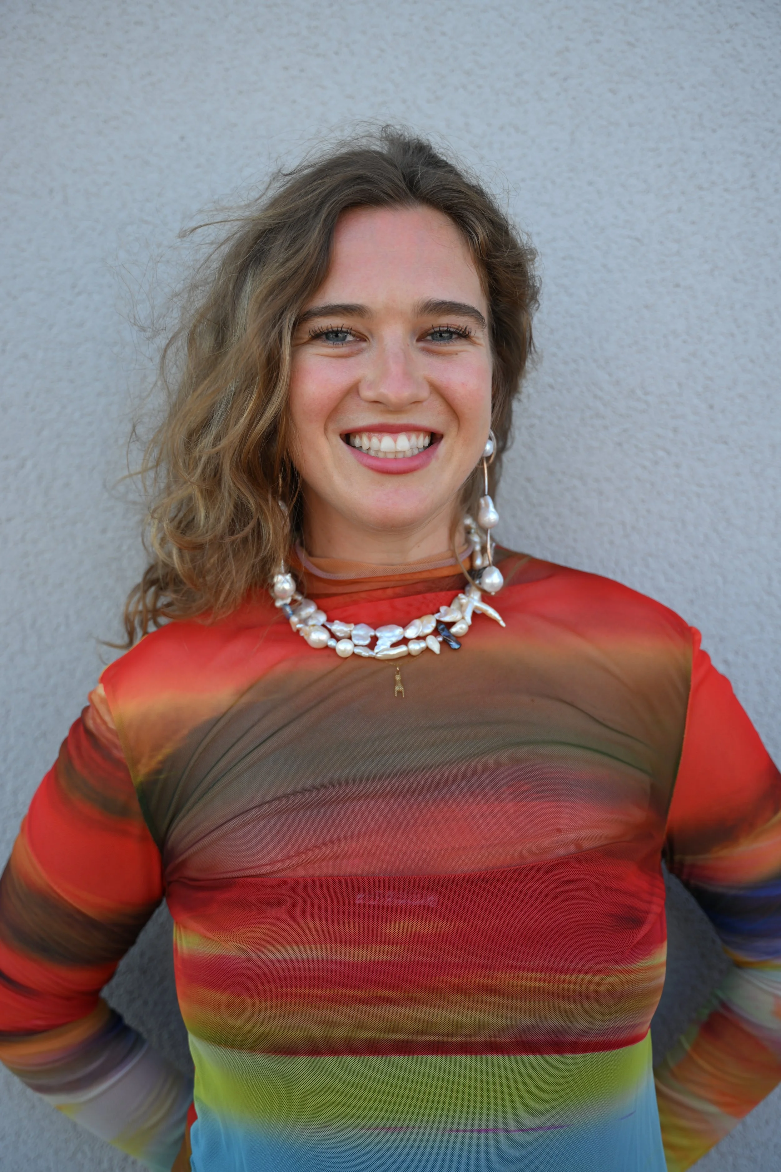 A woman with curly hair smiling, wearing a colorful, patterned dress and a pearl necklace, standing against a plain wall.
