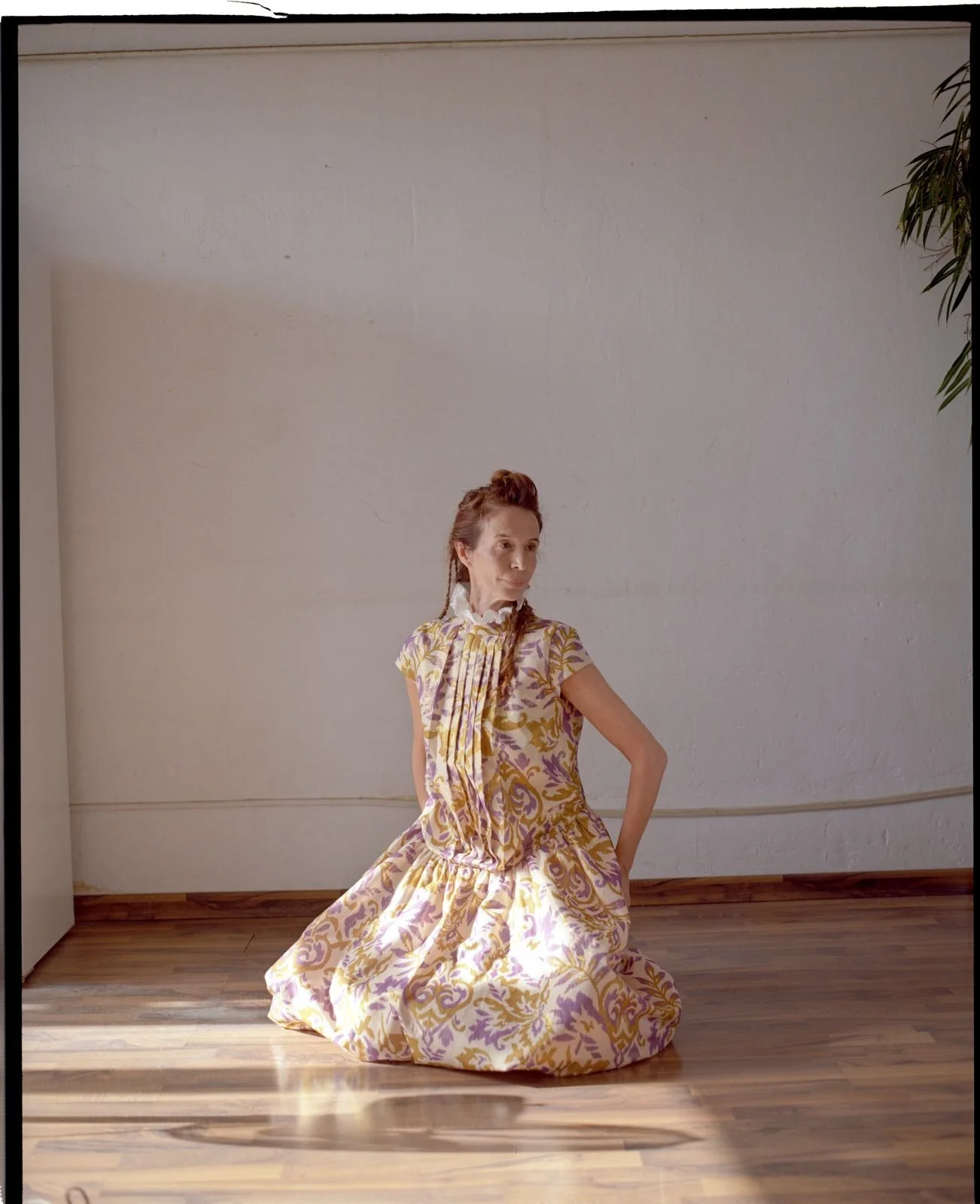 A woman dressed in a vintage floral dress sits on a wooden floor against a plain white wall, with a plant partially visible on the right side of the image.
