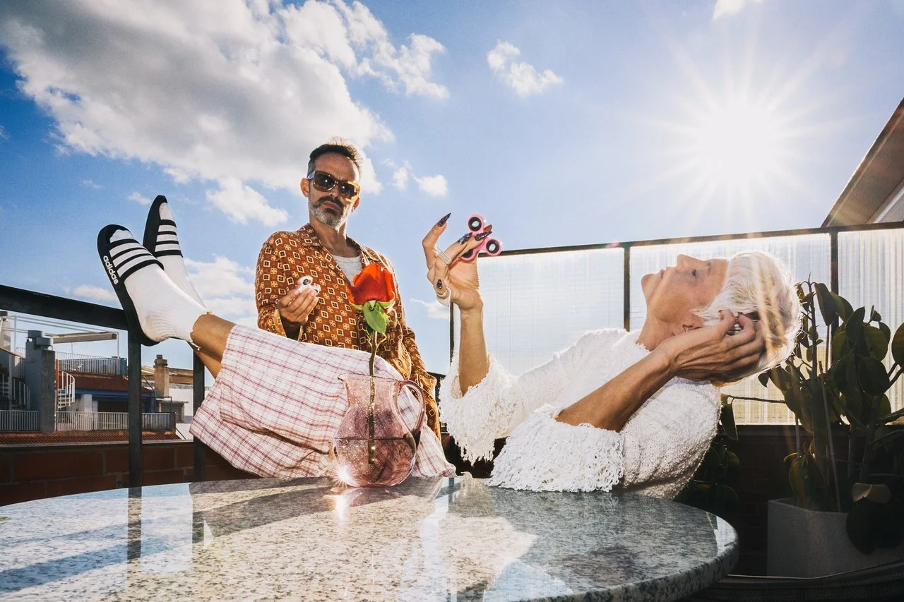 A man and a woman on a rooftop terrace with a glass railing during daytime with sunlight. The woman, with gray hair, is reclining at a round table, wearing a white lace dress, listening to music with headphones. The man, standing, has a beard and mus