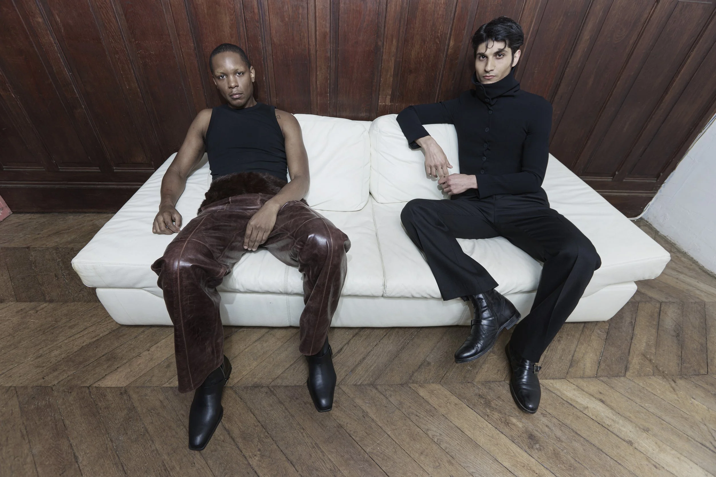 Two individuals sitting on a white couch against a wooden wall and hardwood floor. One person has darker skin, short hair, and is wearing a black sleeveless top, brown leather pants, and black boots. The other has light skin, dark hair styled up, and