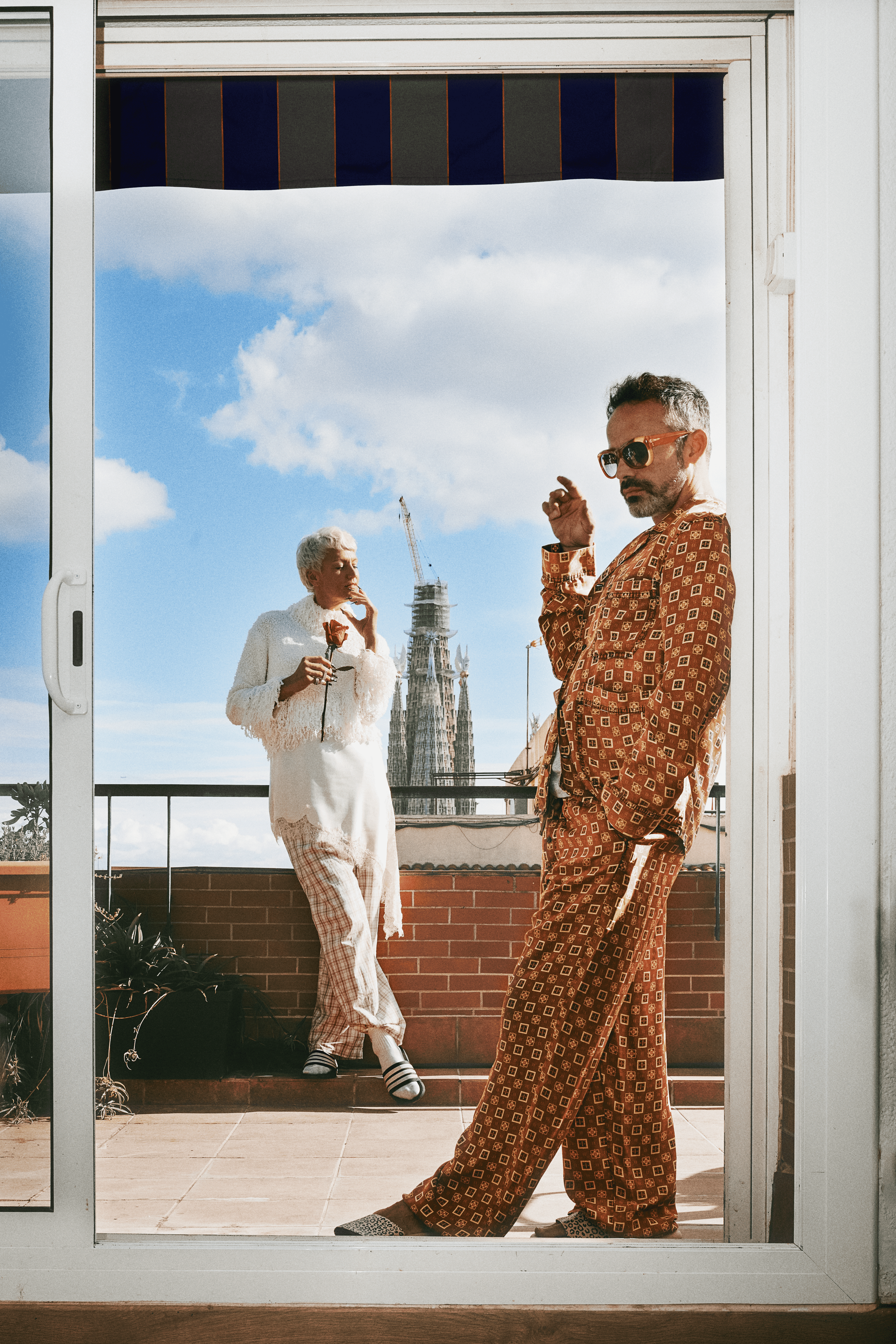 Two people on a balcony dressed in vintage pajamas and robes, with the Sagrada Família church visible in the background under a partly cloudy sky.
