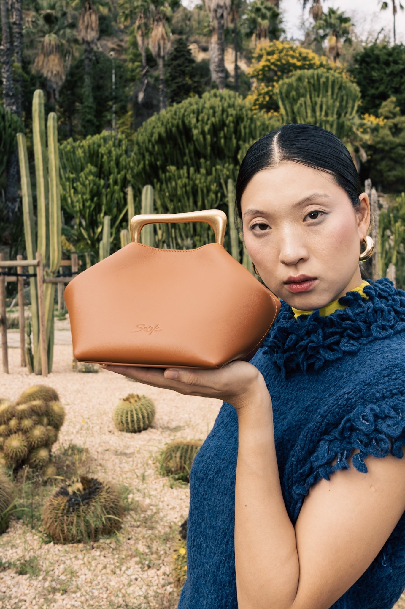 A woman with black hair in a blue textured shirt holds a tan handbag outdoors with cacti and green bushes in the background.