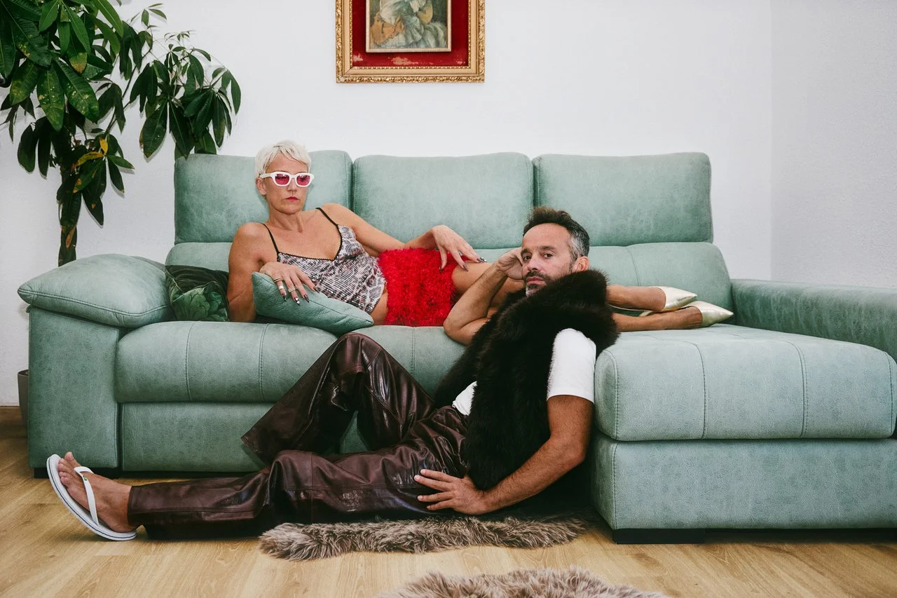 An elderly woman with white hair, wearing pink sunglasses, a patterned top, and red furry shorts, lying on a green sofa. A man with short dark hair, wearing a white T-shirt, burgundy leather pants, and white flip-flops, sitting on the floor beside th