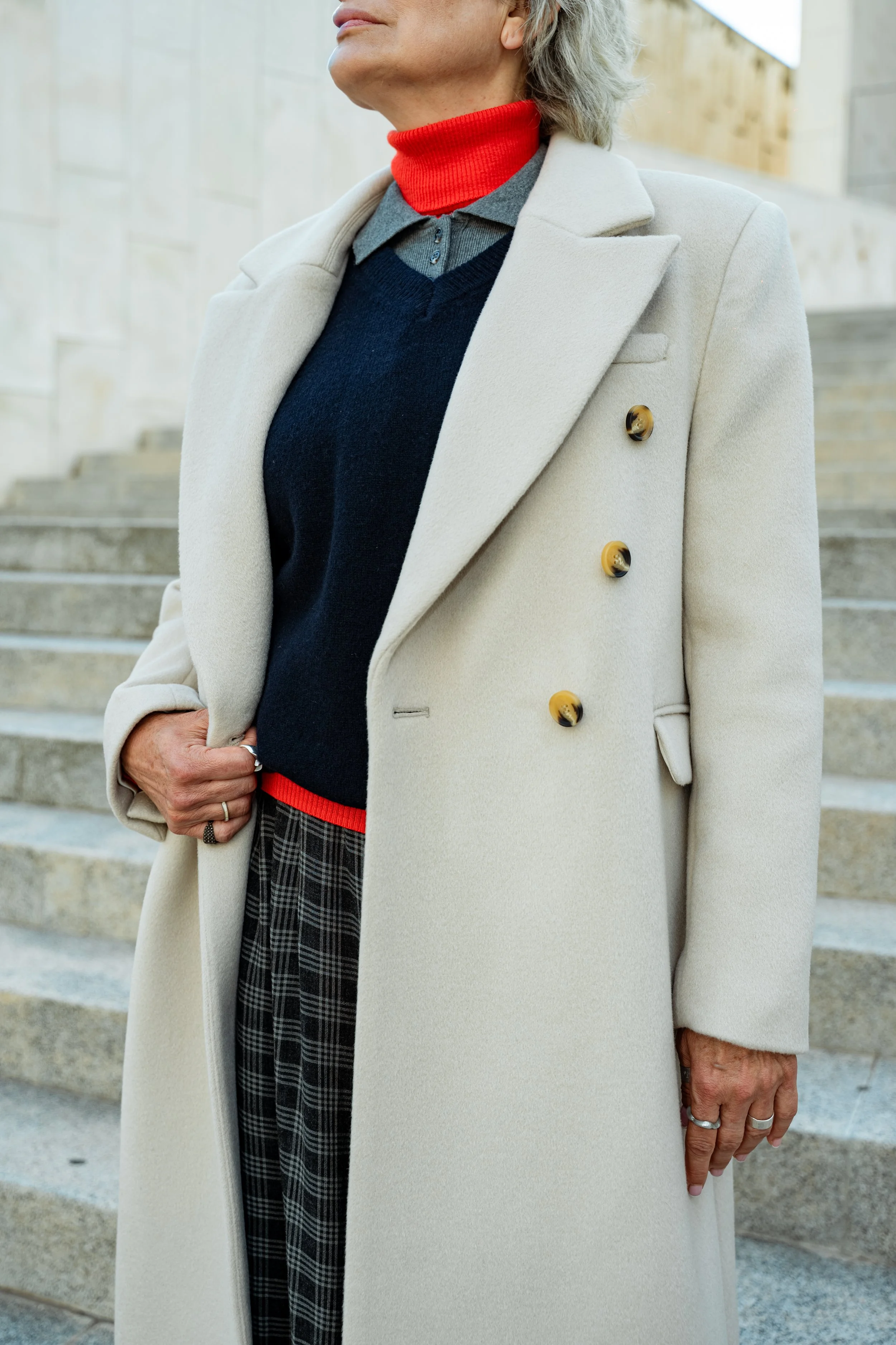 Close-up of a person wearing a light-colored overcoat, red turtleneck, dark sweater, gray shirt, plaid pants, standing on steps outside.