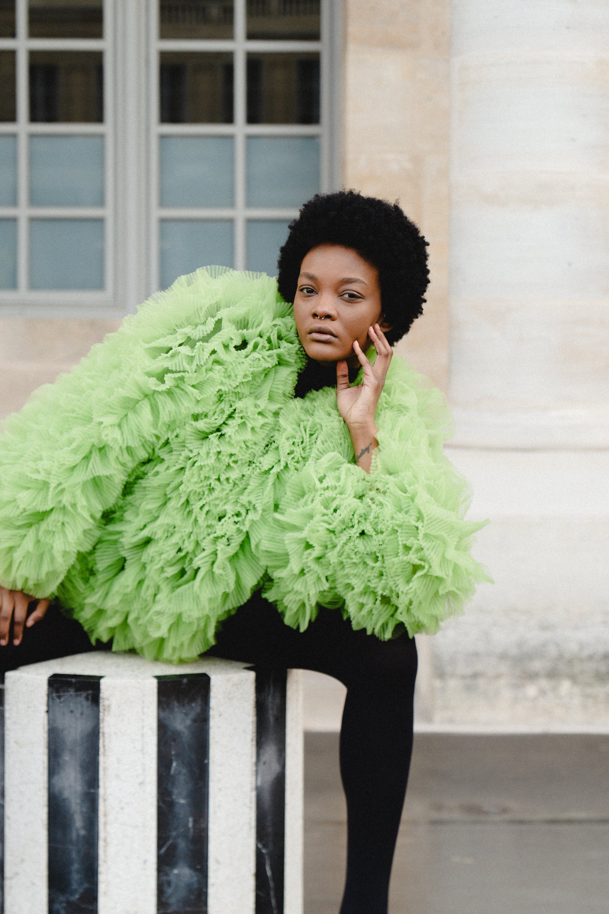 A woman with dark skin and an afro hairstyle, wearing a bright green textured jacket, is sitting outdoors on a black and white rectangular block near a building with a large window, posing with her hand touching her face.