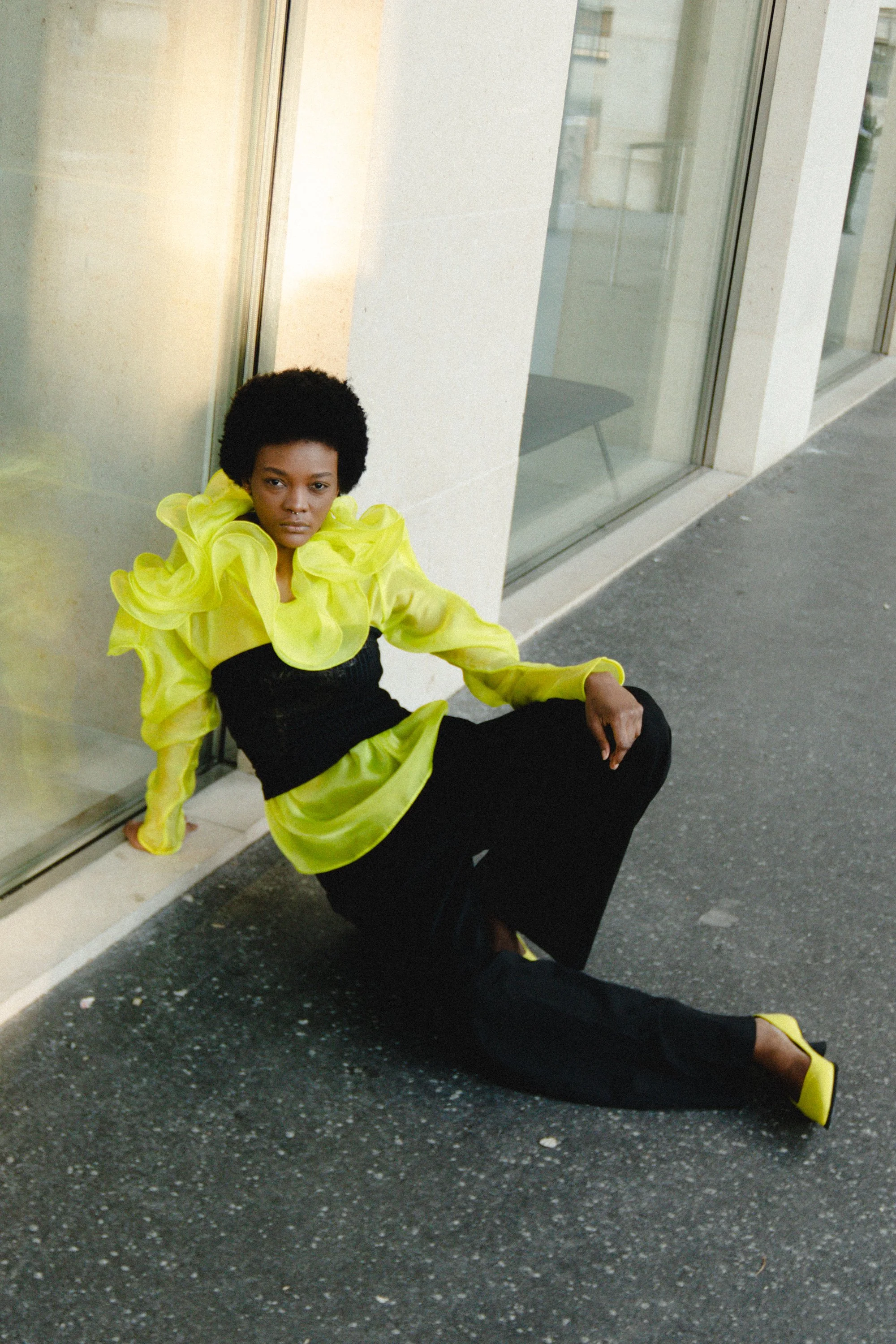 A woman with dark skin and short curly black hair sitting on the pavement outside a building with large glass windows, wearing a bright yellow ruffled blouse, black pants, and yellow shoes.