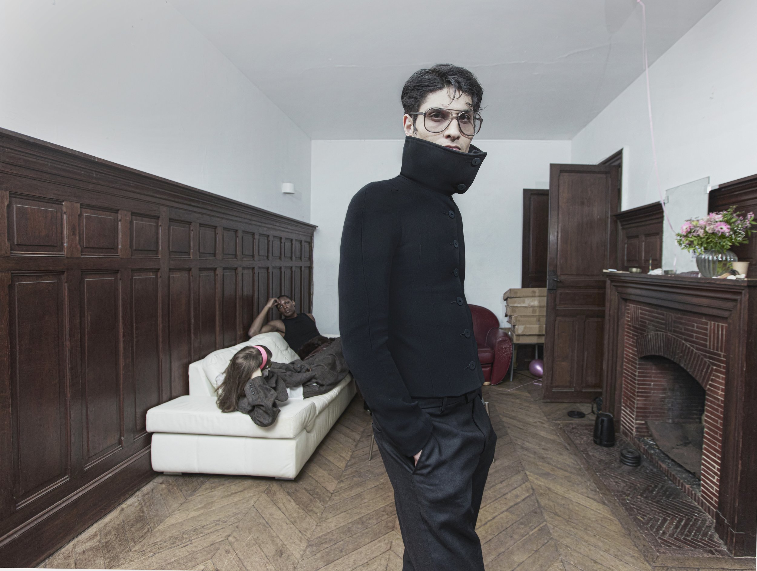 A man in black clothing and glasses standing in a room with raised eyebrows. In the background, a woman lies on a white couch with a young girl sitting next to her.