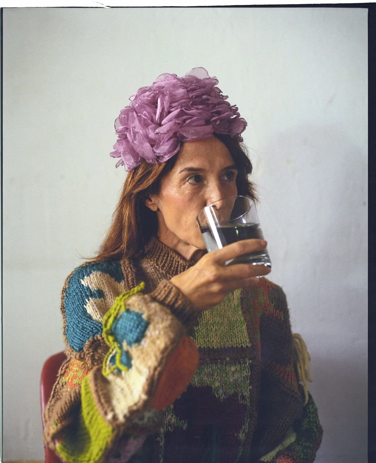 A woman with a purple flower hat drinking water from a clear glass, wearing a colorful, patchwork sweater