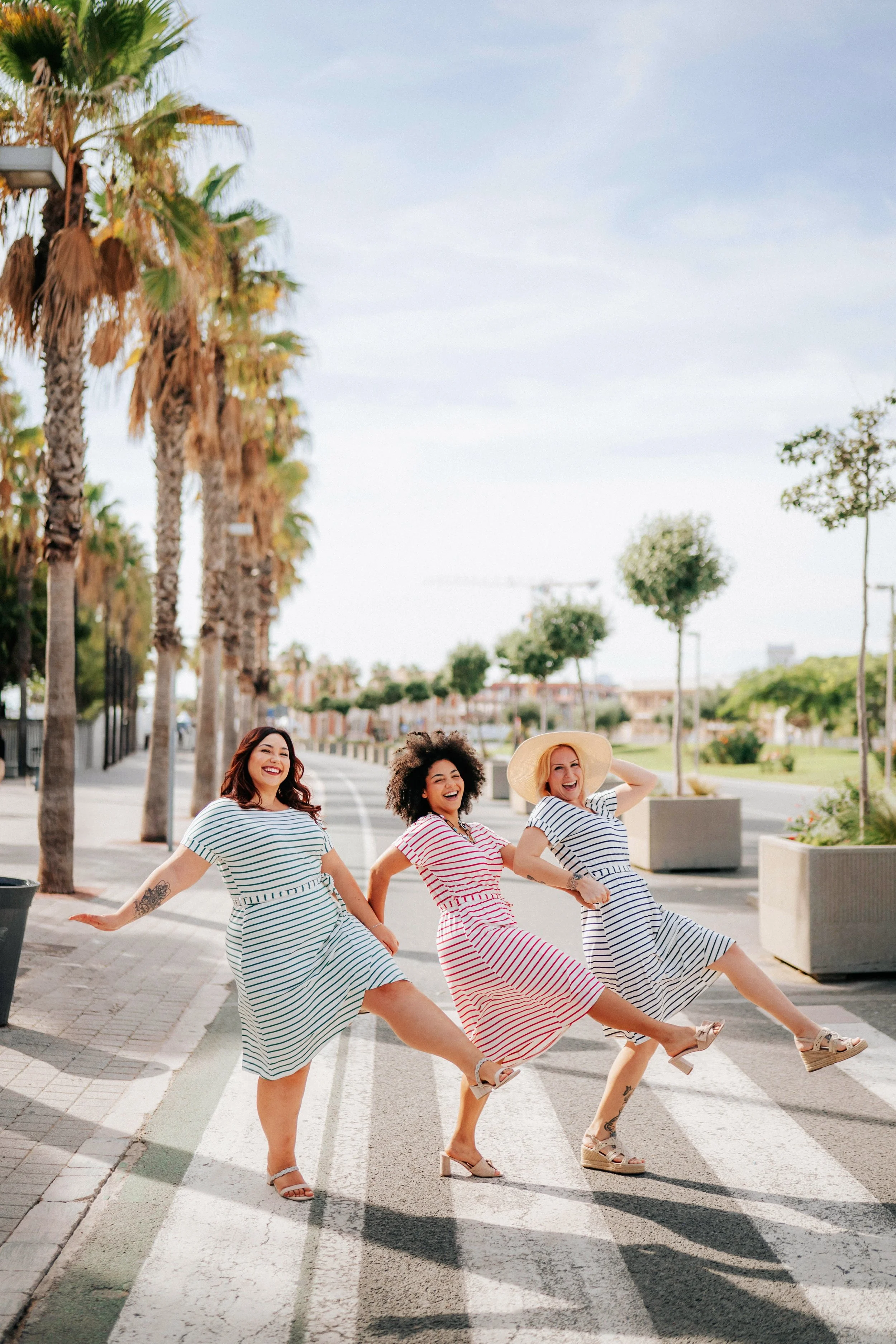 Three women in striped dresses are holding hands and lifting their legs, smiling and walking on a crosswalk with palm trees in the background on a sunny day.