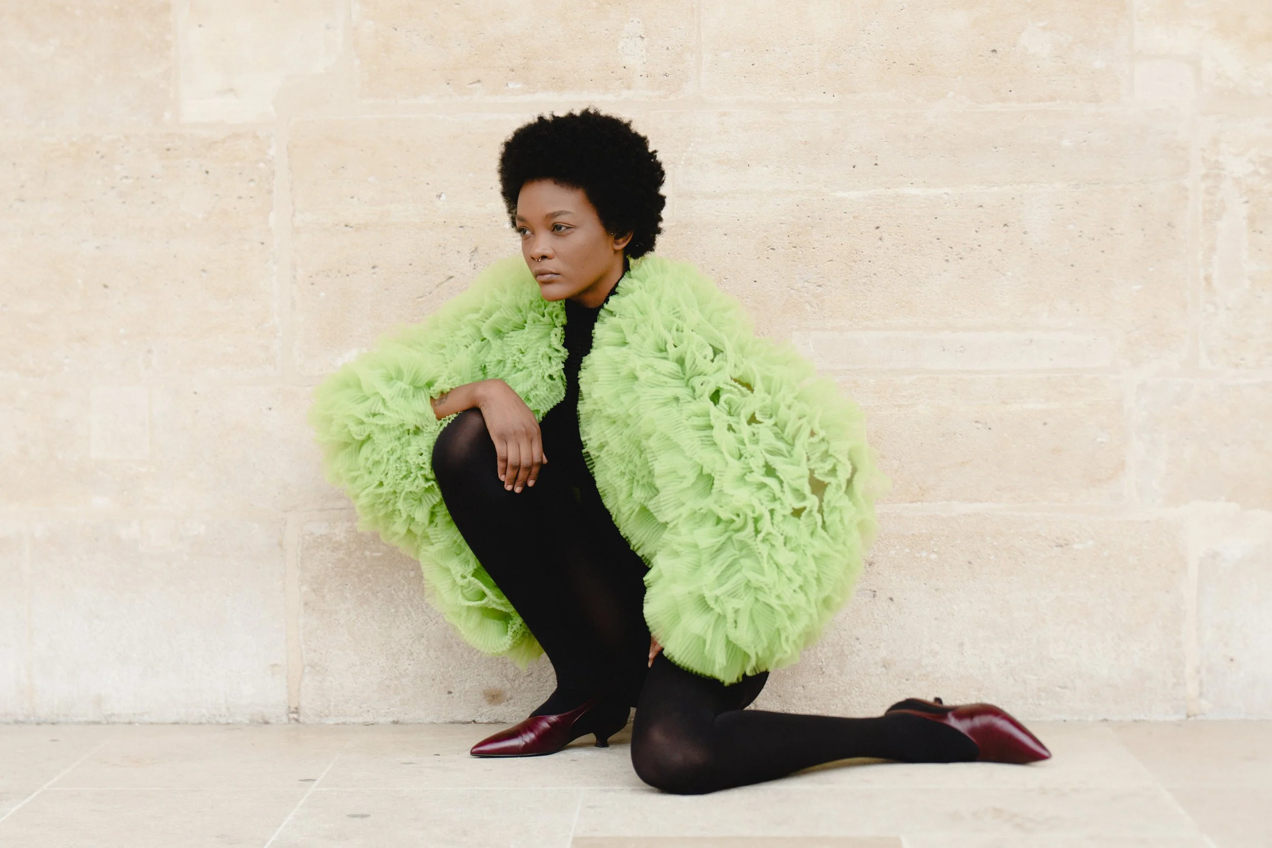 A woman with short curly hair is squatting against a beige stone wall, wearing a bright green ruffled jacket, black tights, and red pointed shoes.