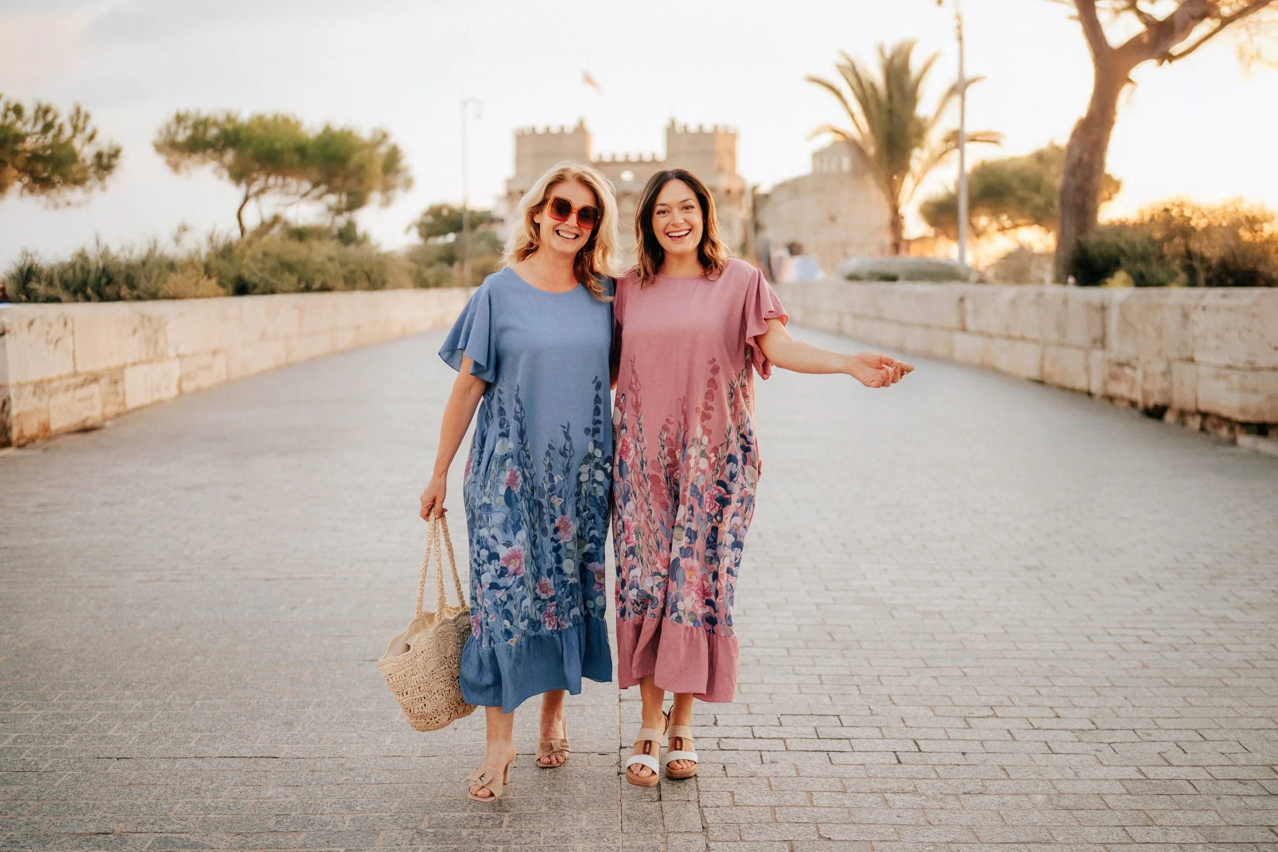 Two women in colorful dresses walking together on a sunny street, smiling and enjoying each other's company, with a castle and trees in the background.