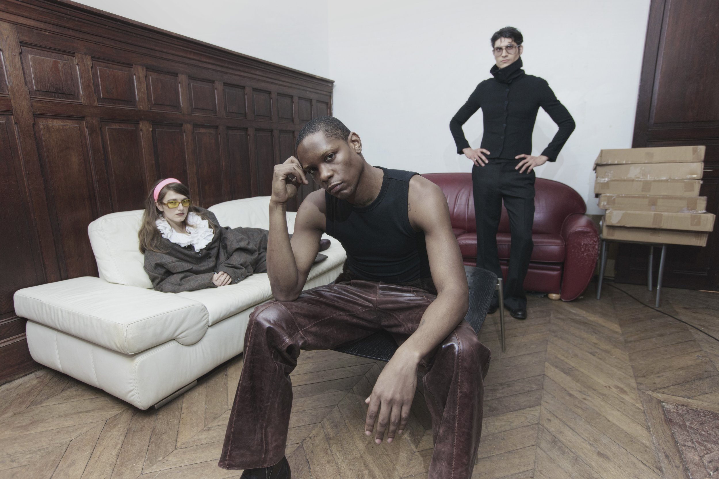 Three models posing in an indoor setting with wooden wall paneling and a wooden floor. The male model in the foreground is sitting on a black chair, wearing a black sleeveless top and brown leather pants, with his hand on his head. The female model i