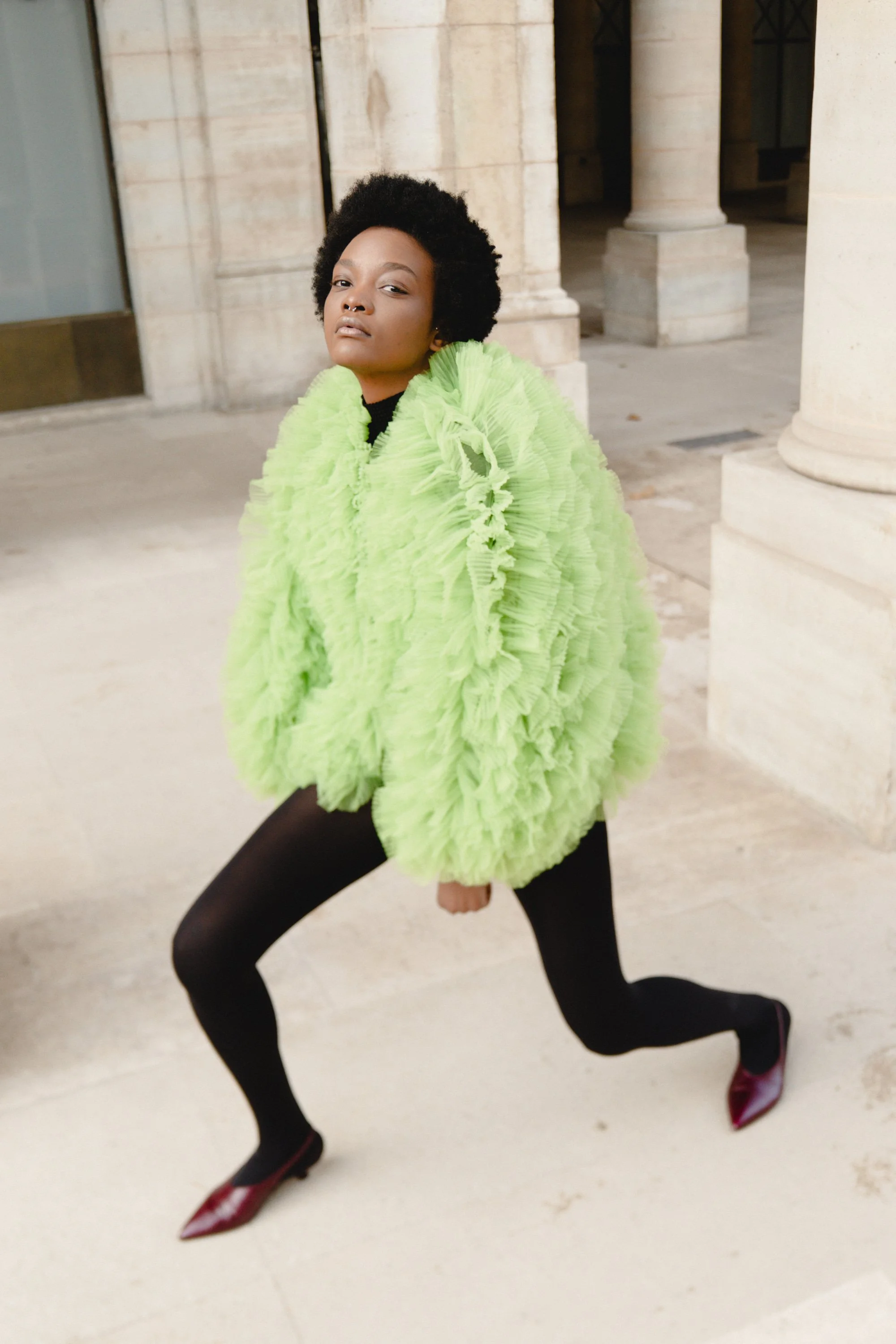 A woman in a bright green ruffled coat, black tights, and red heels posing confidently outside near stone columns.