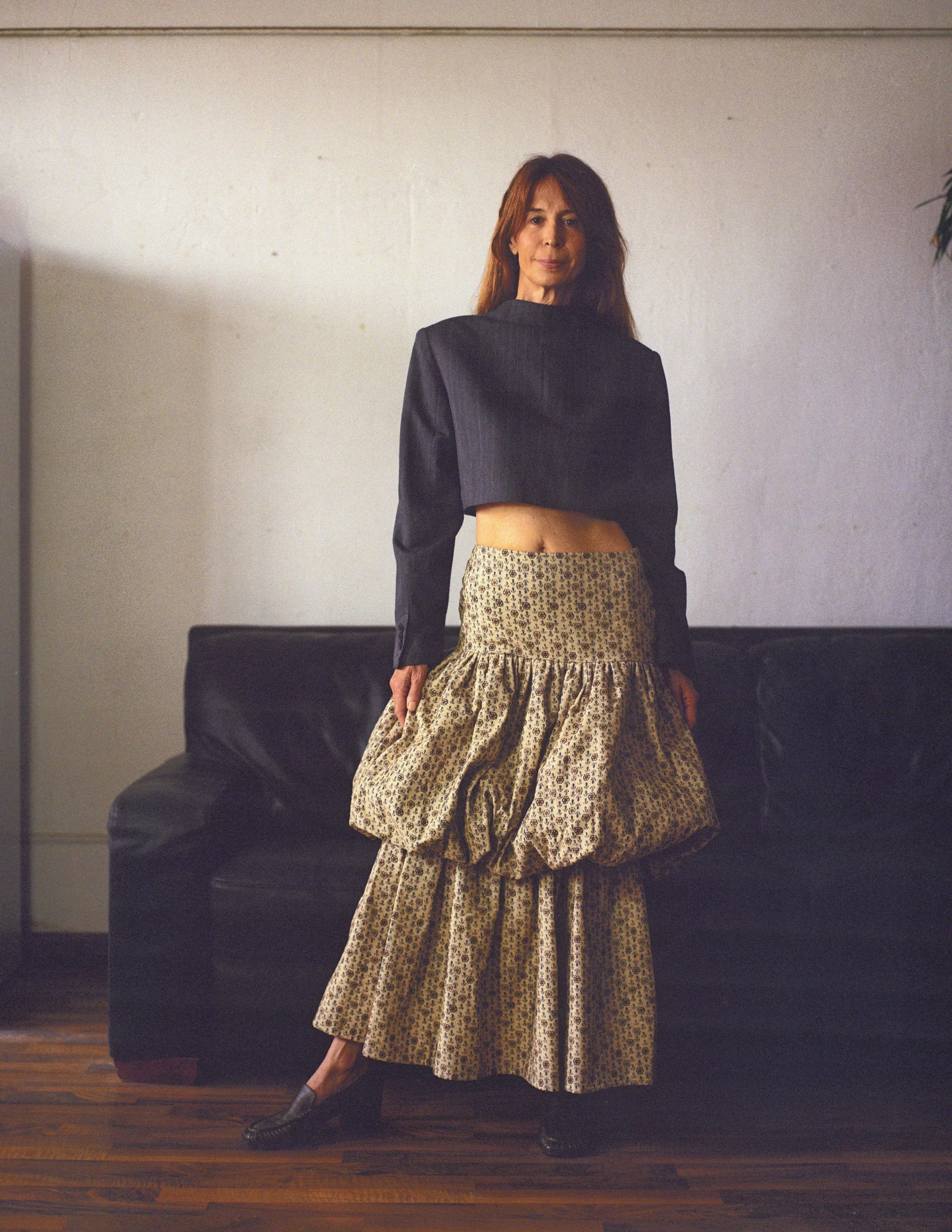 Woman standing in front of a black leather couch, wearing a black long-sleeve top and a high-waisted, layered, animal print skirt with a gathered, voluminous design.