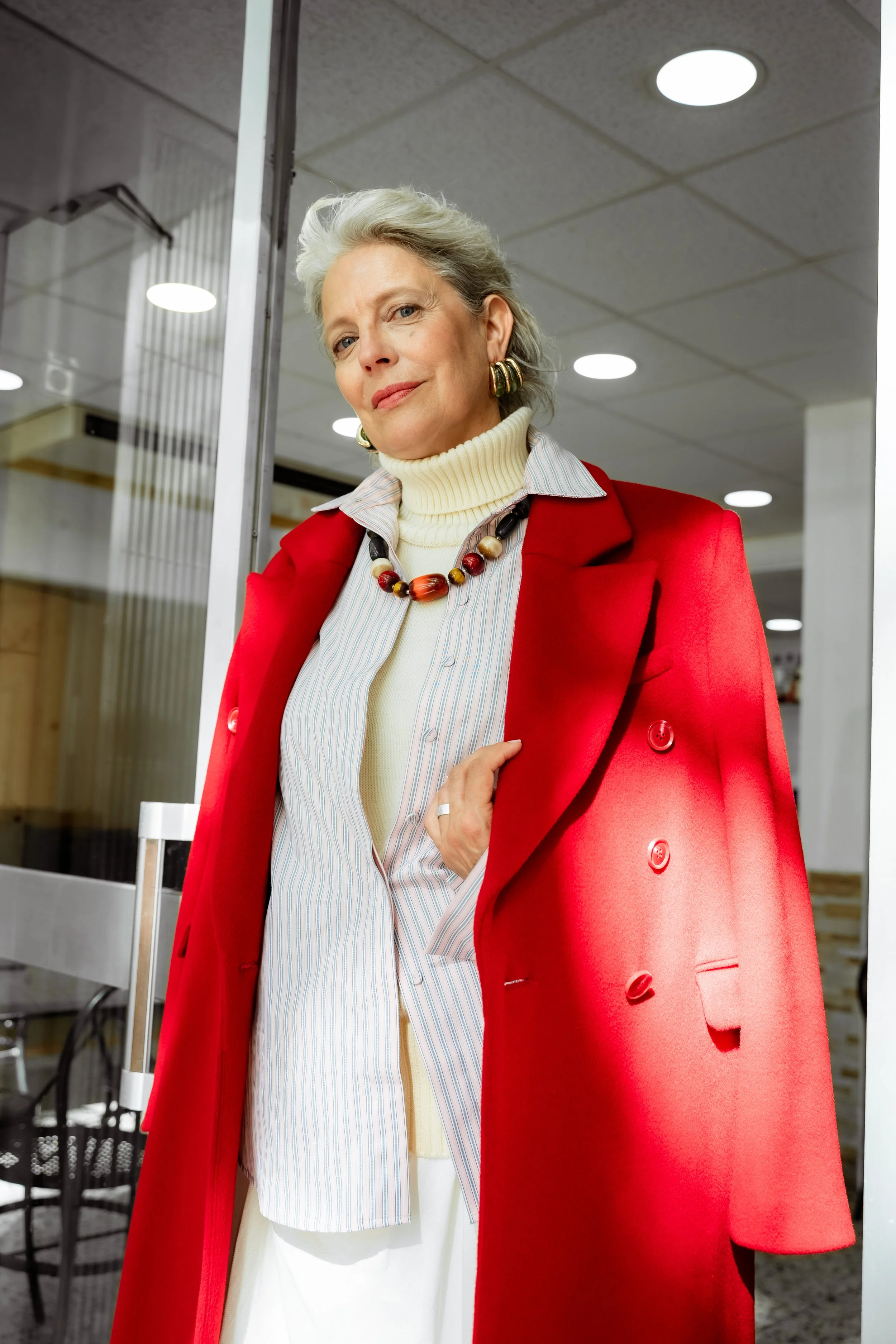 An older woman with gray hair wearing a red coat, striped shirt, cream turtleneck, and colorful necklace. She is standing indoors, leaning slightly, with a confident expression.