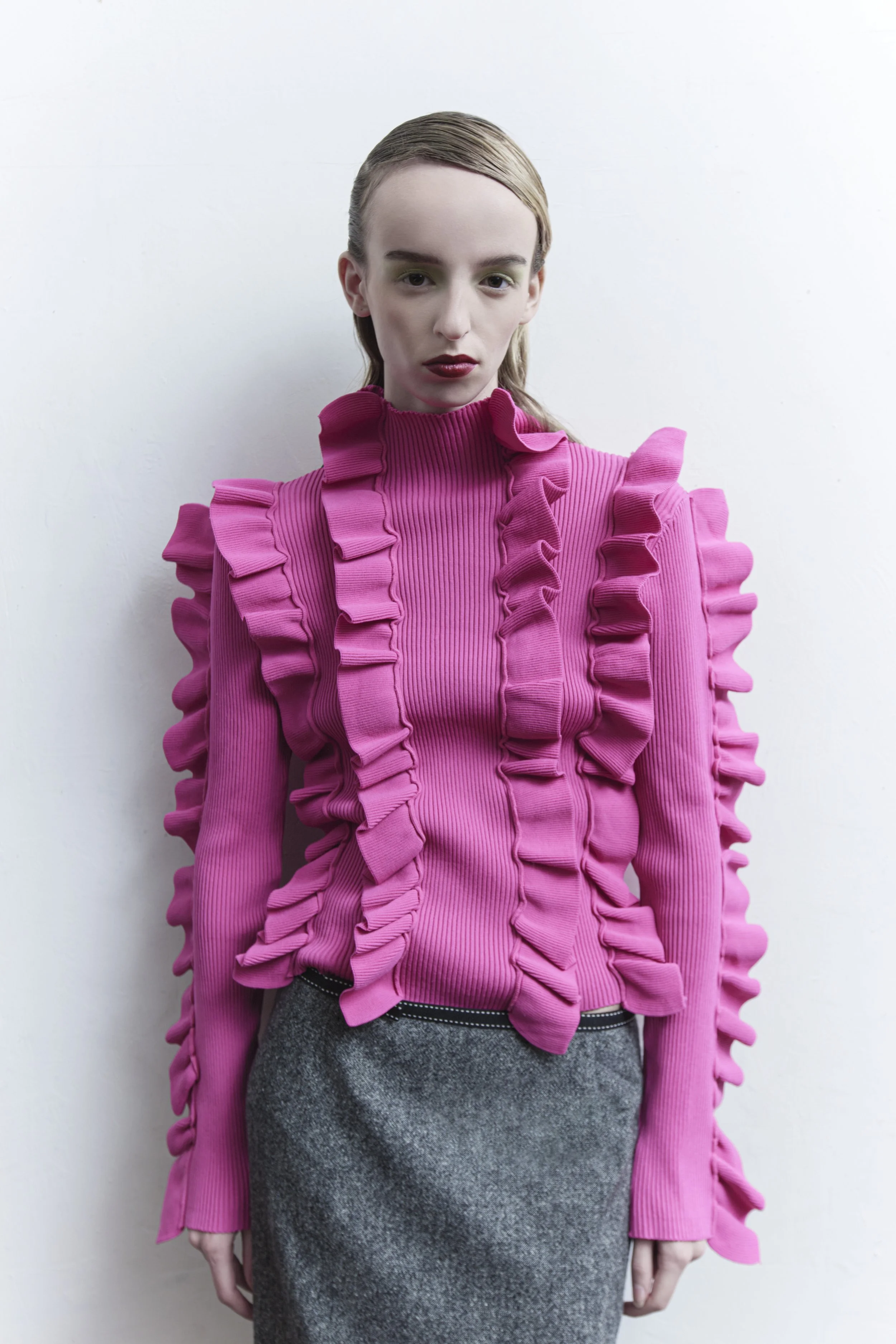 A woman standing against a white wall wearing a bright pink ruffled long-sleeve top and a gray skirt.