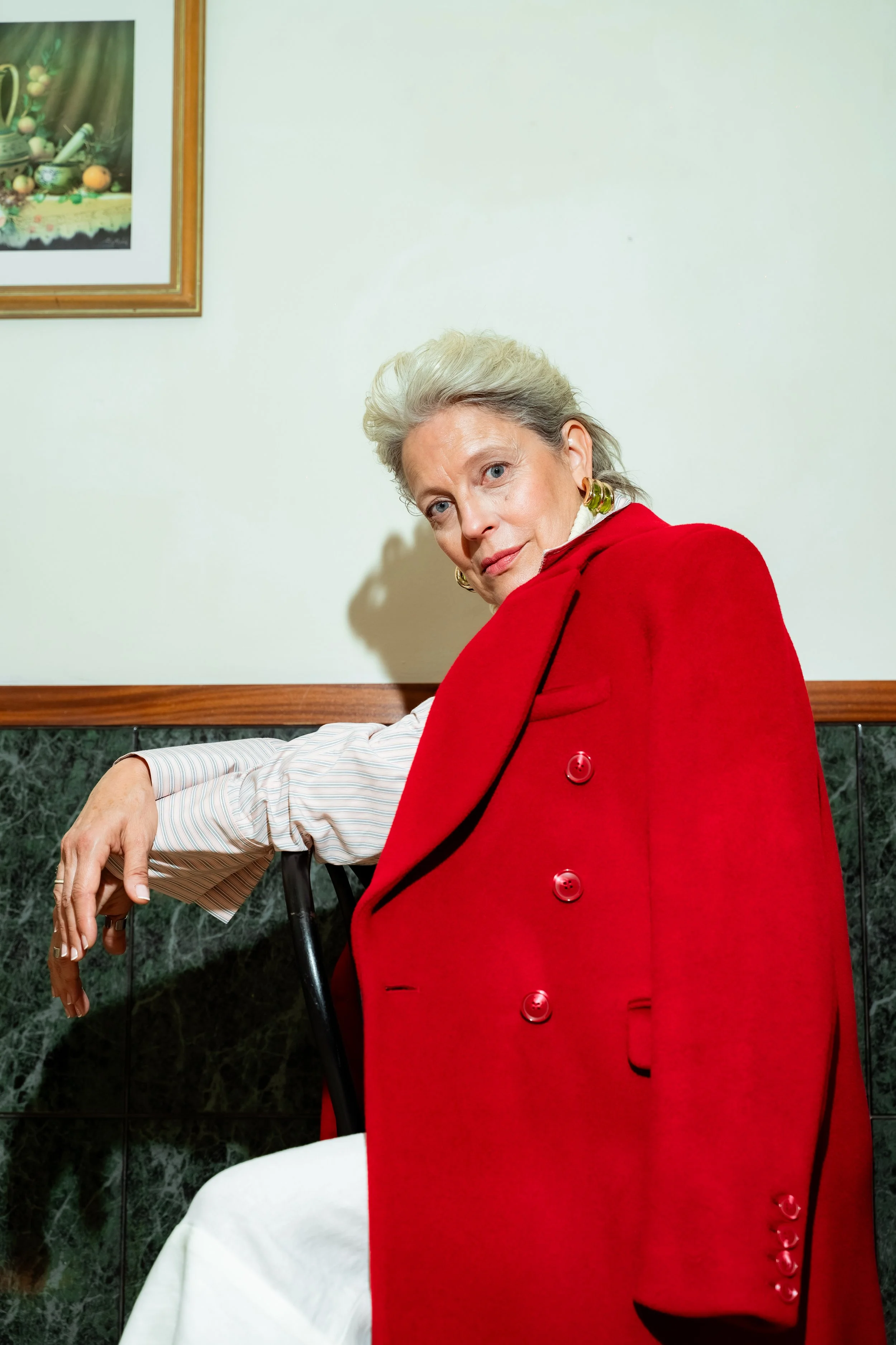 An elegant older woman with gray hair dressed in a red coat and earrings, sitting on a black chair with her arm resting on the back, looking at the camera in a room with a light-colored wall, a marble wainscot, and a framed painting.