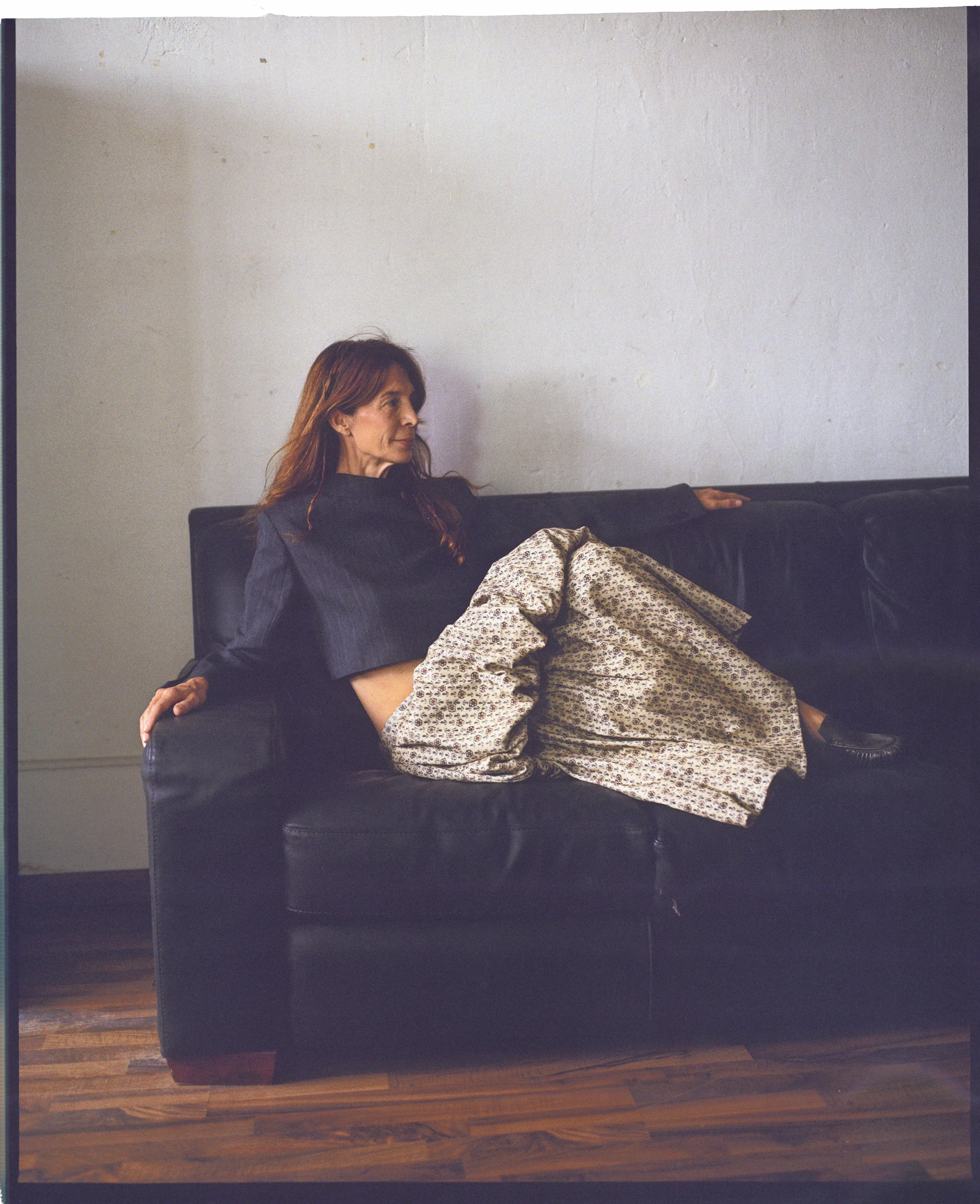 A woman with red hair lounging on a black leather couch against a plain white wall, wearing a dark top and patterned loose pants.