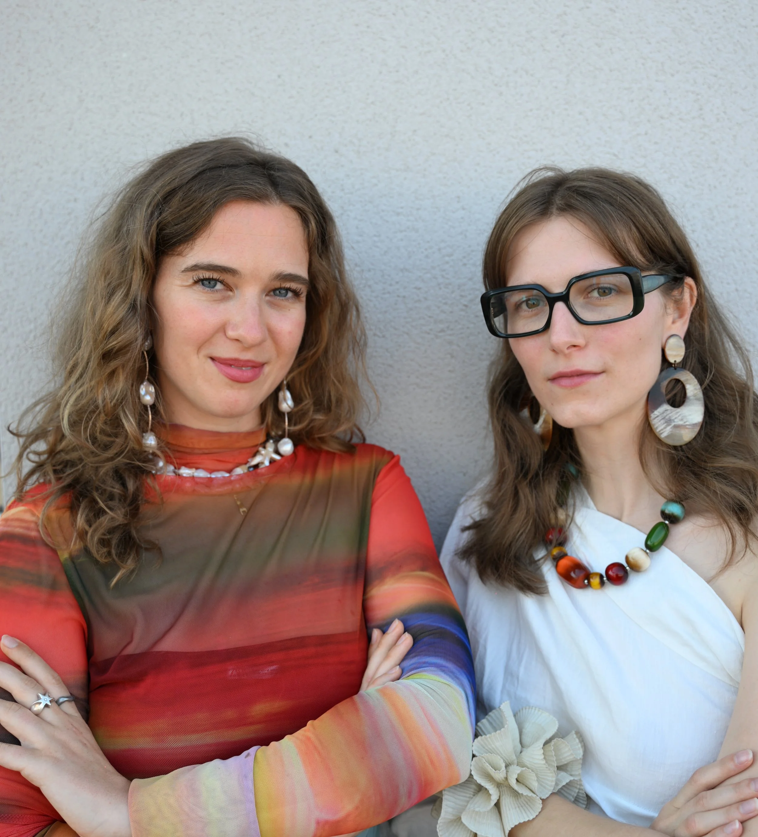 Two women standing against a plain wall, with arms crossed, looking at the camera. One has shoulder-length curly brown hair, wearing a colorful long-sleeve top and jewelry. The other has shoulder-length wavy brown hair, glasses, and is dressed in a white top with a large bow detail, wearing colorful jewelry.