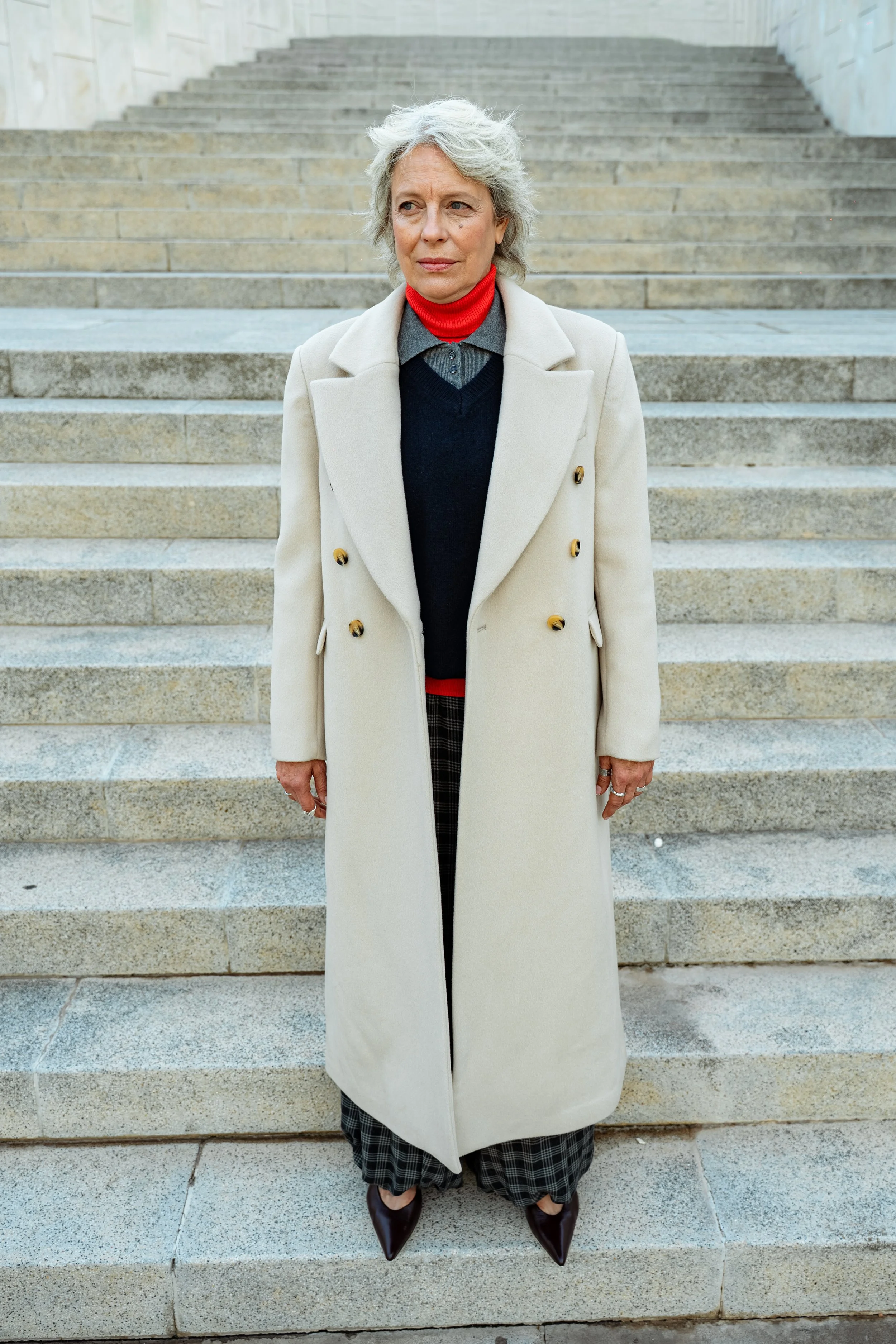 A woman with gray hair standing on outdoor stone steps, wearing a long beige coat, red turtleneck, gray sweater, plaid pants, and dark shoes.