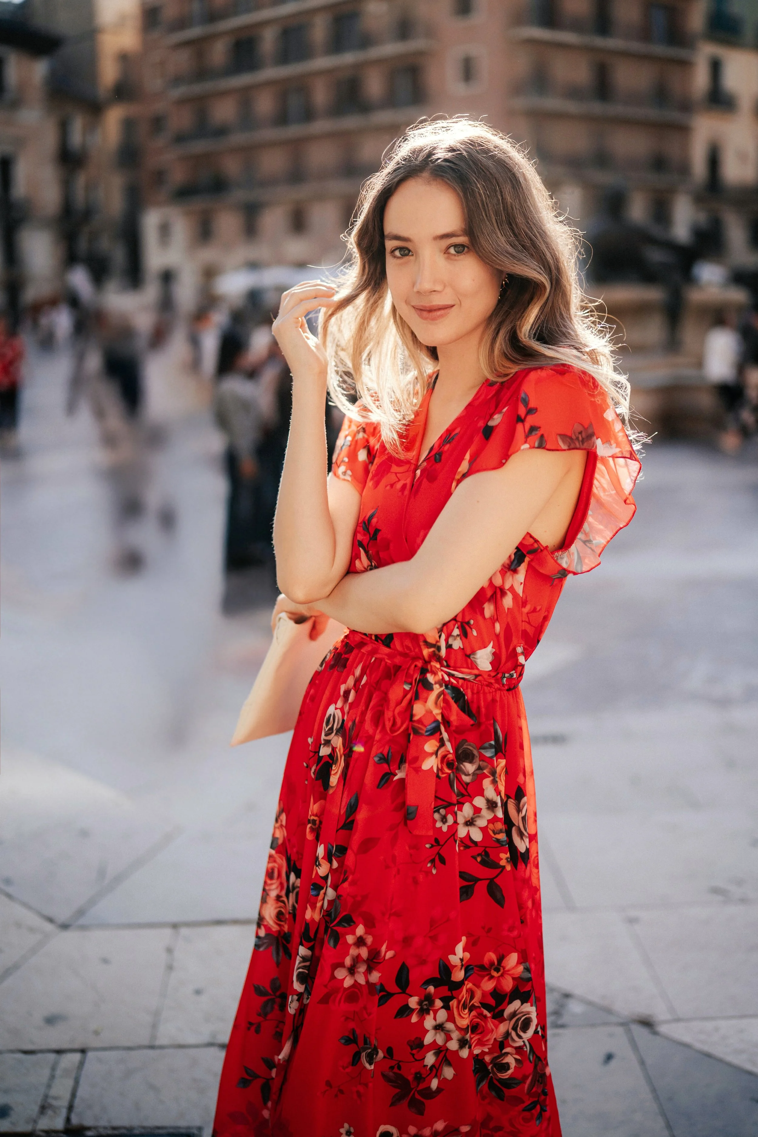 A young woman with wavy brown hair, wearing a red floral dress, standing outdoors on a city street with blurred pedestrians and buildings in the background.