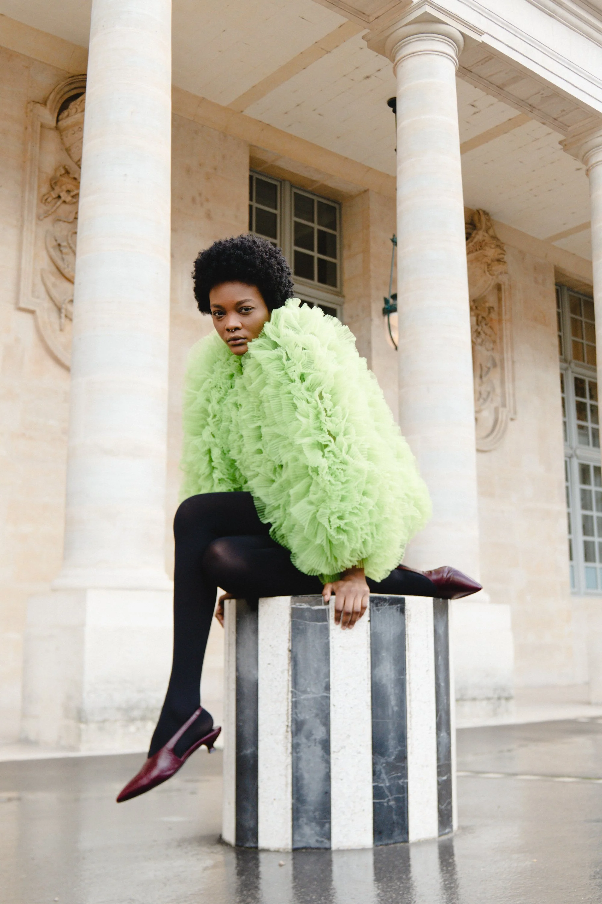 A woman with short curly black hair wearing a bright green ruffled jacket, black tights, and burgundy high heels, sits on a marble pedestal outside a historic building with large columns and detailed stone carvings.