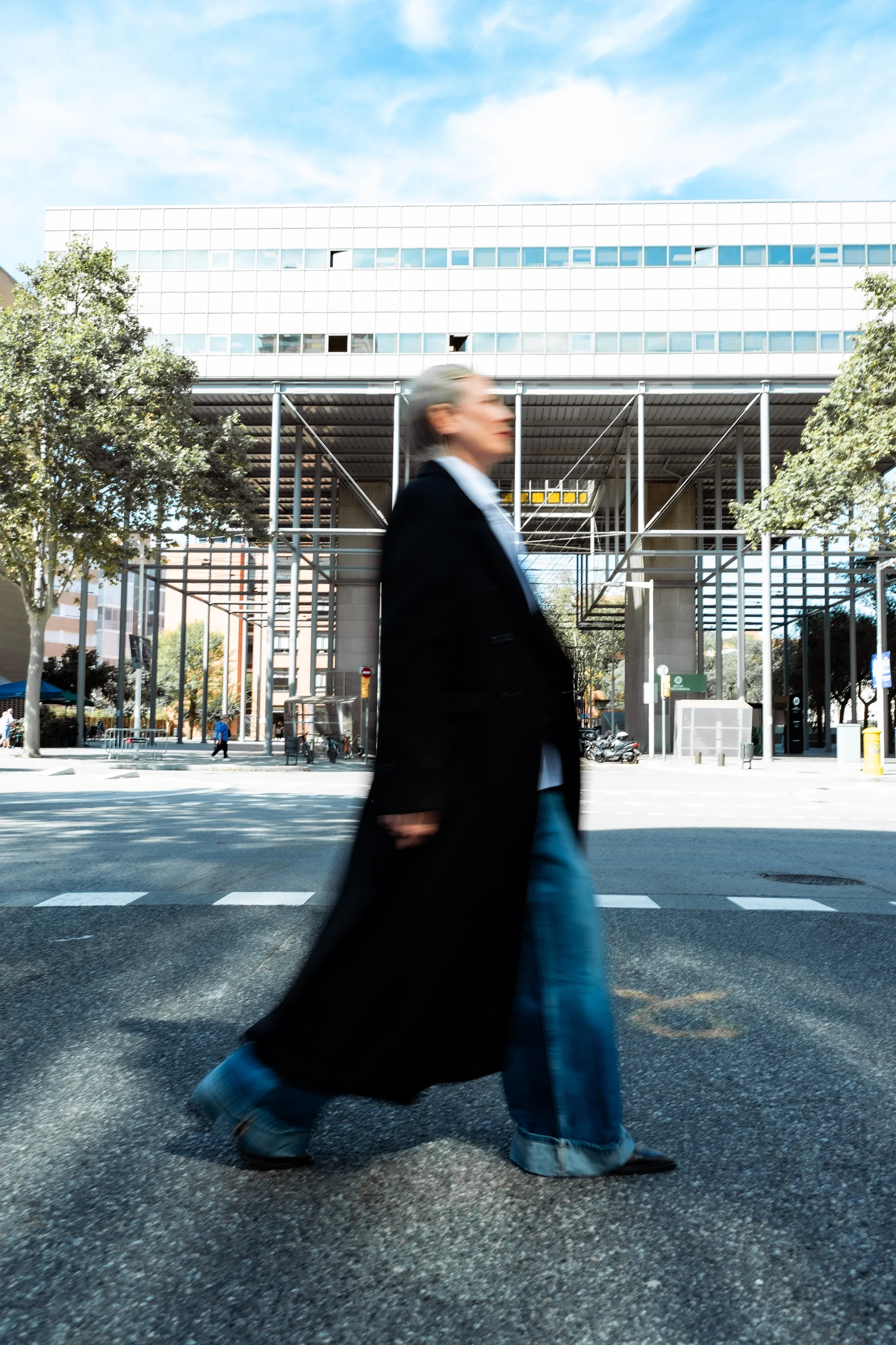 Blurred woman walking across an urban crosswalk in front of a modern building with trees on the sidewalk.
