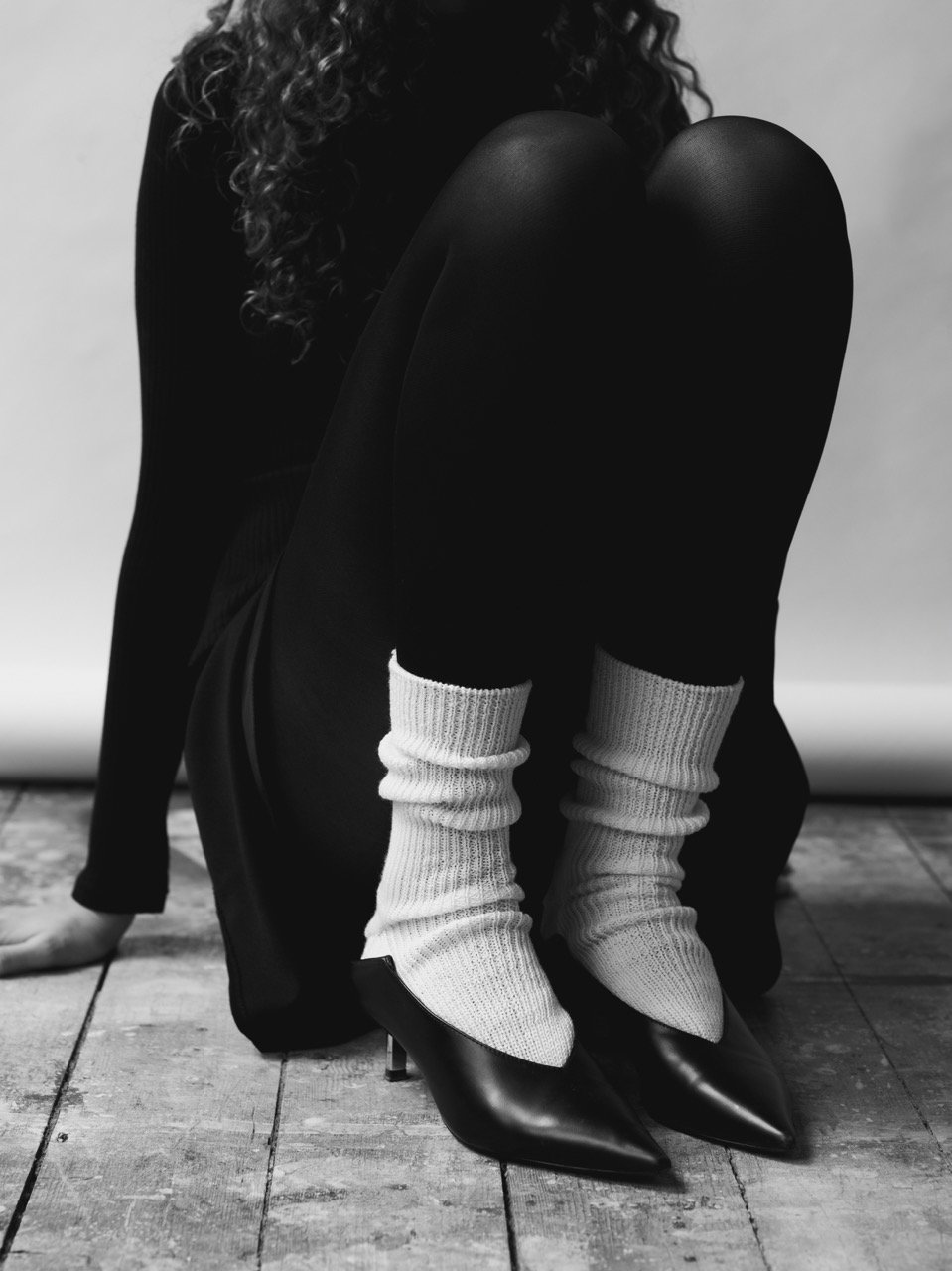 A woman with curly hair sitting on the floor, wearing black tights, pointed black high heels with stiletto heels, cream-colored socks scrunched down, and a black skirt or dress.