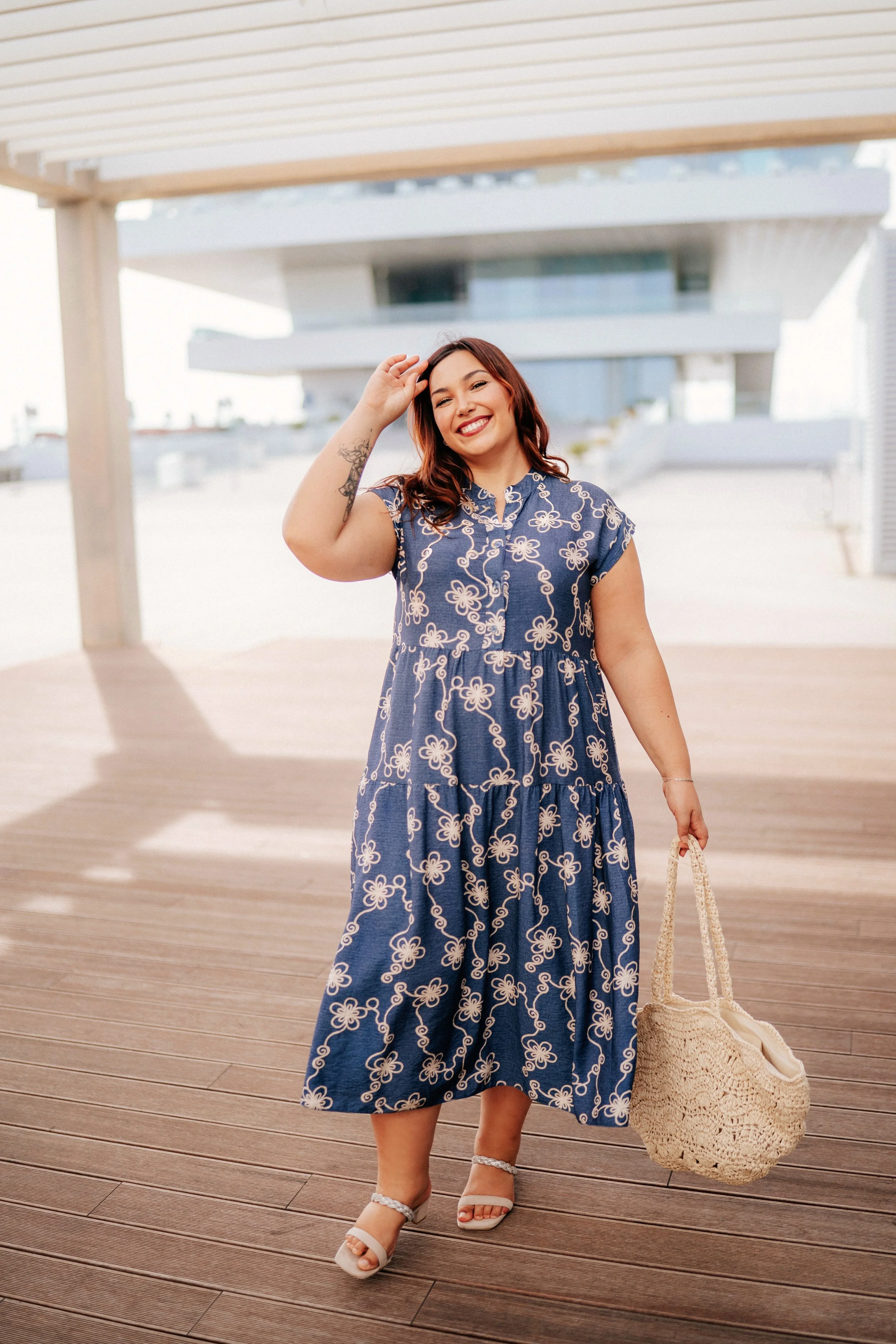 A woman in a blue floral dress standing on a wooden deck, smiling and holding a woven straw bag, with a modern building in the background.