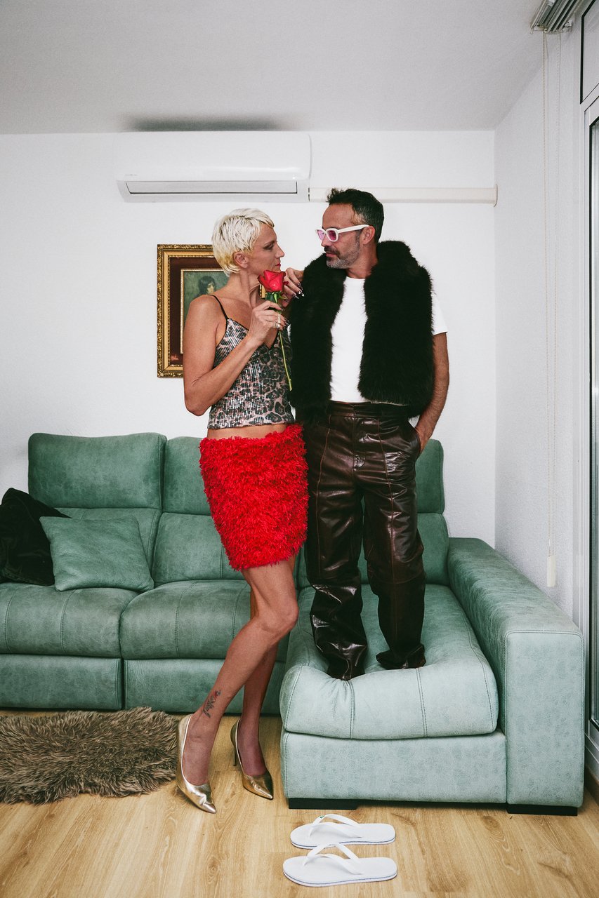 A woman with short blonde hair and a man with dark hair and glasses standing on a green sectional sofa in a modern living room. The woman is holding a red rose close to her face, wearing a leopard print top, red textured skirt, and metallic high heel