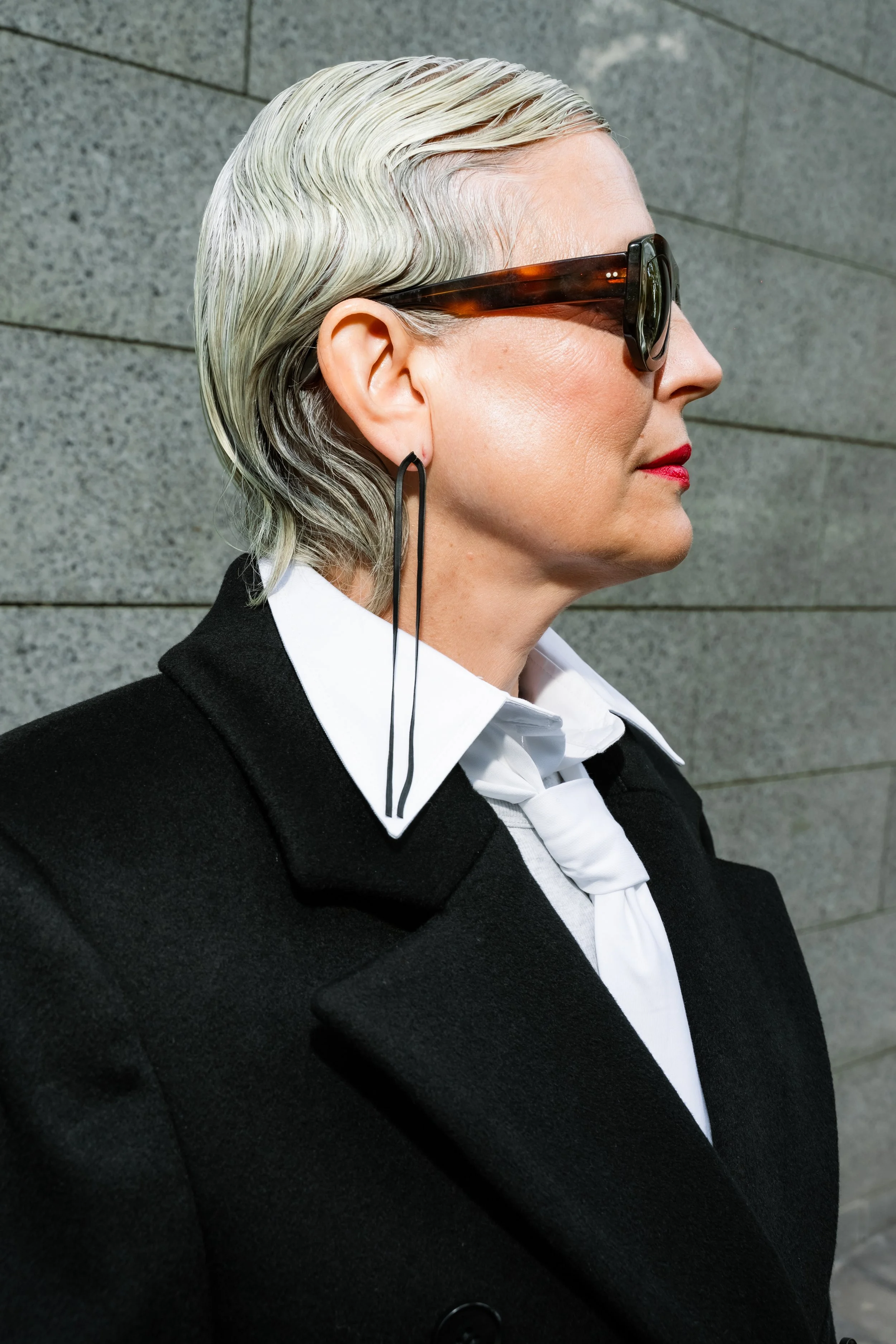 Side profile of a woman wearing large sunglasses, a black coat, white shirt, and a white scarf, against a gray tiled wall.