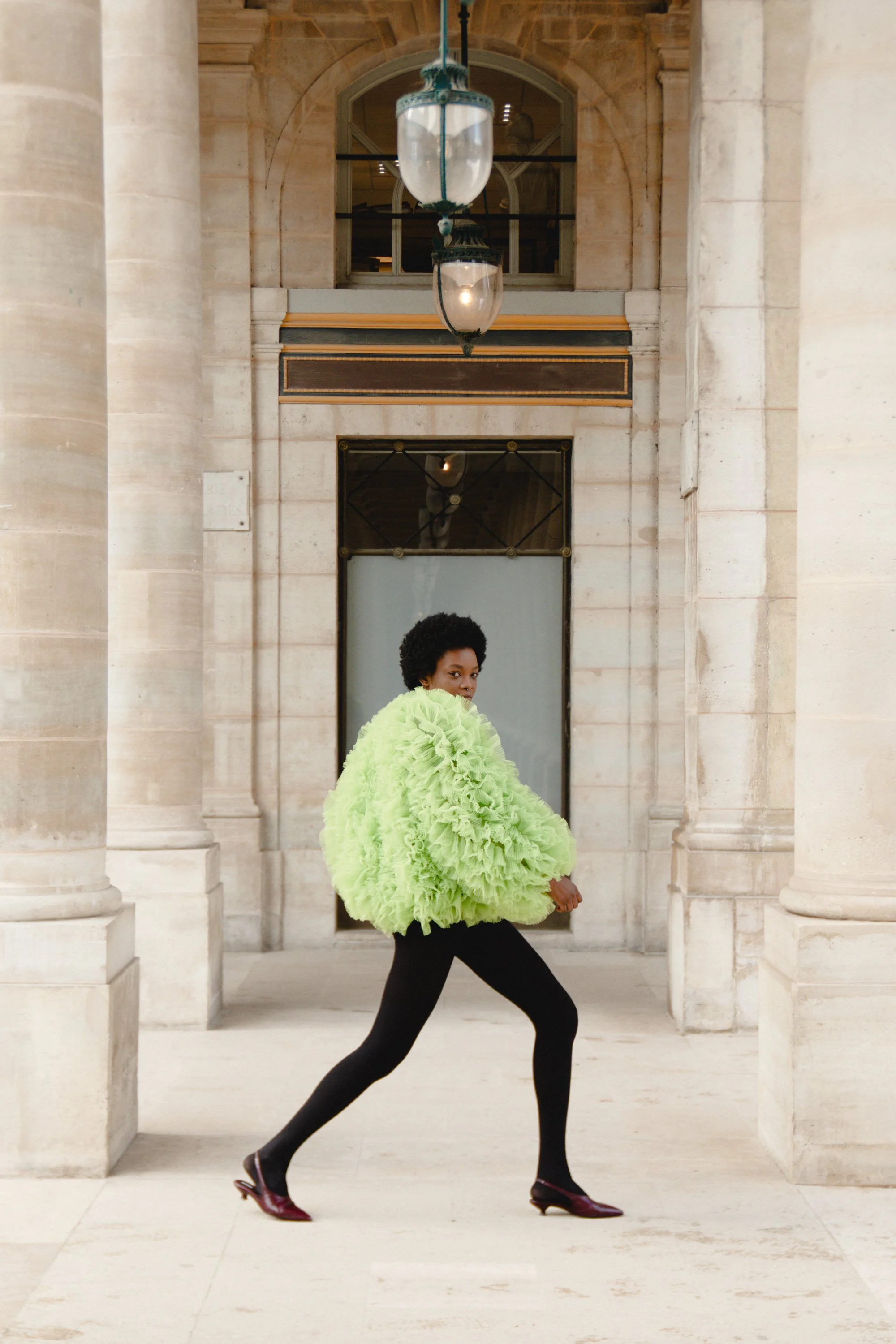 A woman with short curly hair wearing a bright lime green fluffy jacket, black leggings, and burgundy heels, poses confidently in front of a stone building with large arches and columns, under a hanging lantern.