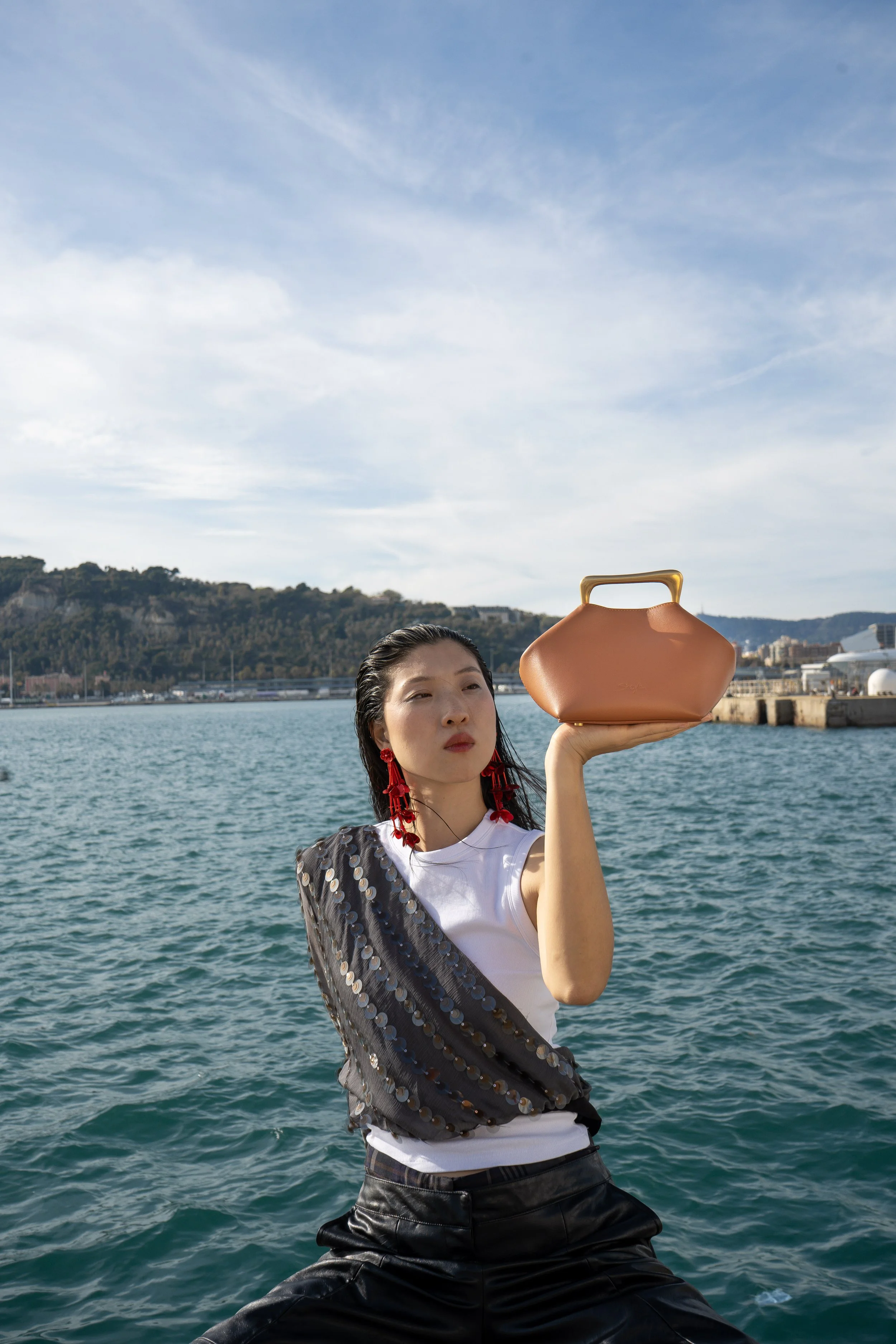 A woman standing outdoors near water, holding a peach-colored handbag up in the air with her right hand, wearing a white sleeveless top, black leather pants, and red statement earrings, with a scenic harbor and hills in the background.